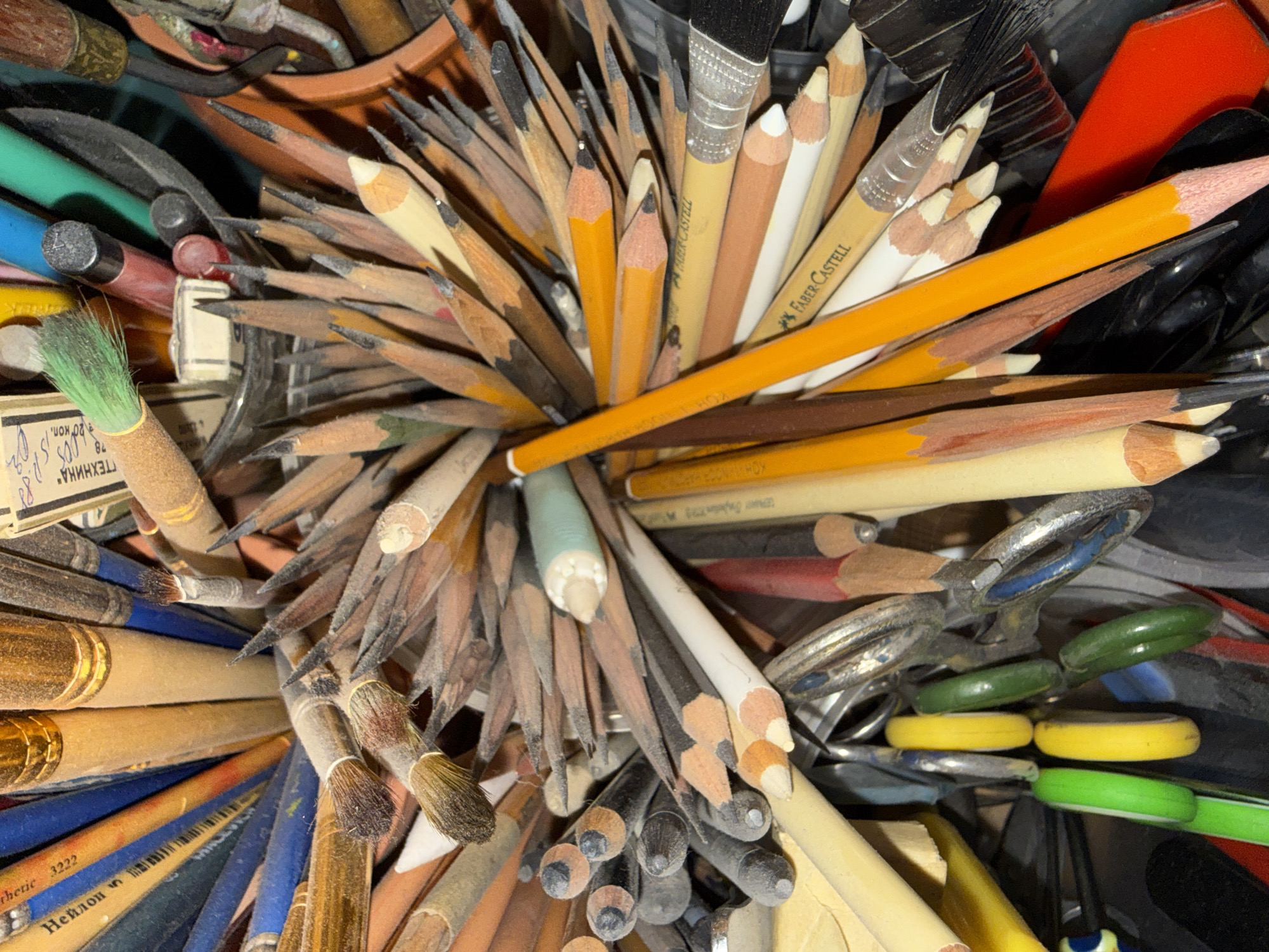 This is a close-up, overhead shot of a large collection of art supplies, primarily pencils and paintbrushes, densely packed into a container. The lighting is somewhat dim and artificial, highlighting dust particles on the surfaces of the objects, suggesting the collection has been in use for some time. The pencils are of various colors and degrees of sharpness, with some appearing well-worn and others recently sharpened. The brushes also vary in size and bristle type, some with wooden handles and others with metallic ferrules. Interspersed among the pencils and brushes are other art-related items such as scissors and what appear to be erasers. The background is dark and indistinct, offering no specific environmental context. The image is devoid of any human presence or interaction. The scene appears to be a personal art studio or a storage area for art supplies. There is no information or visual cue to suggest this location is in Votkinsk, Russia.