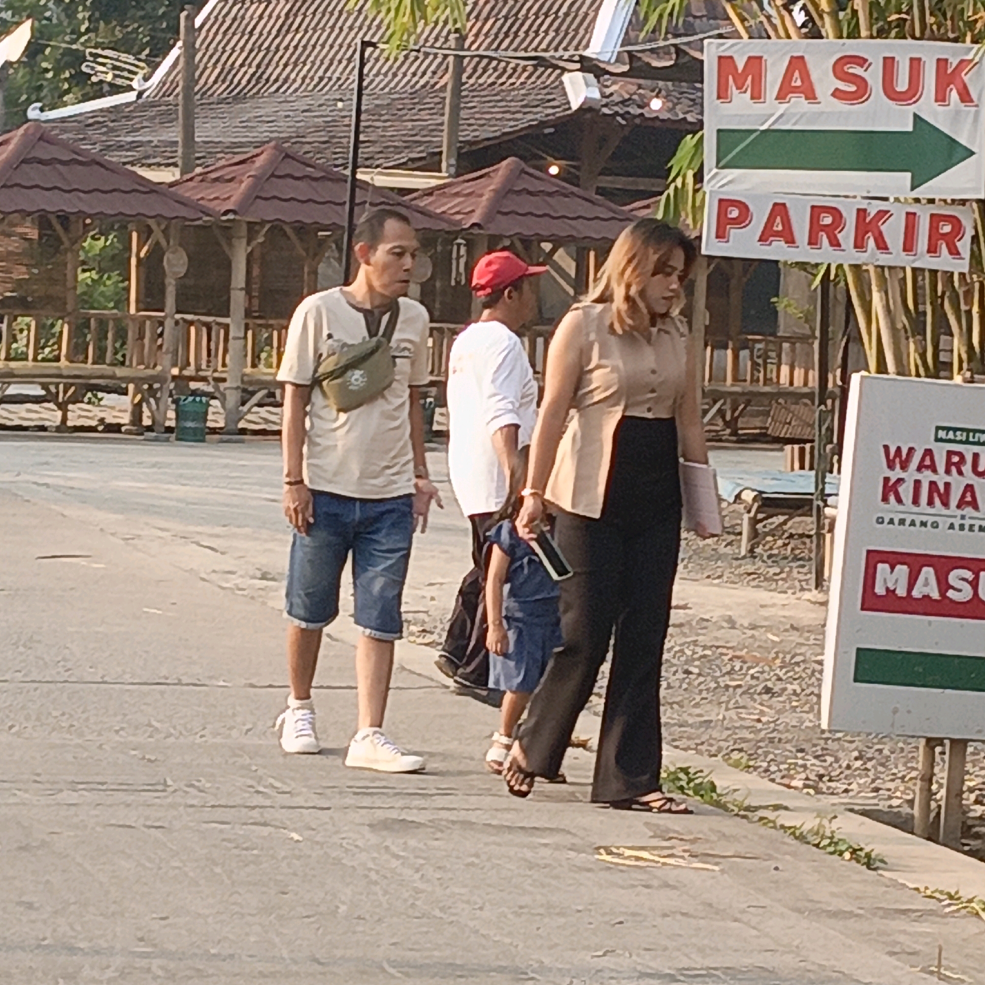 In Candirejo, Indonesia, a group of people walk along a paved path. The foreground shows the legs and lower bodies of several individuals. A man on the left wears denim shorts and white sneakers, with a green crossbody bag over his tan t-shirt. Next to him, a child in a blue dress is held by the hand of a woman wearing dark pants and a light brown button-down shirt. Another person in a white t-shirt walks behind them. In the background, a sign with red lettering reads "MASUK PARKIR" with a green arrow pointing right. Several bamboo structures and thatched-roof gazebos are visible, suggesting an outdoor dining or recreational area. A green trash can is also present. The lighting indicates it is daytime, possibly late afternoon.