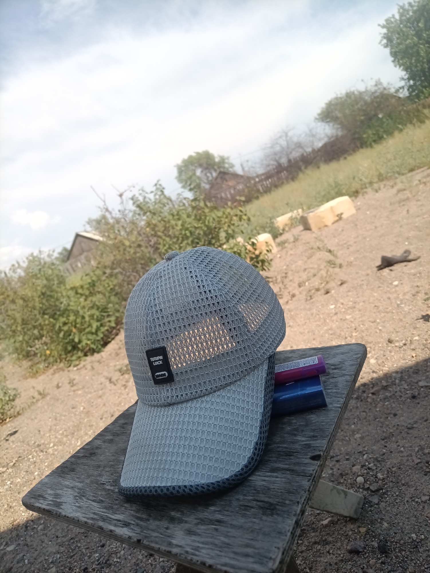 An eye-level, medium shot captures a gray mesh baseball cap resting on a weathered wooden plank in an outdoor setting. The cap is positioned in the foreground, angled to the left, with its brim facing the viewer. Attached to the cap is a black tag with white text that reads "TUTUPAL LUCK" and "MADE". Two lighters, one red and one blue, are partially visible beneath the brim of the cap.
The wooden plank appears to be part of a makeshift table or bench, with a visible support made of concrete. The ground around the plank is covered in dry dirt and small pebbles. In the background, there is dense green shrubbery and trees, and a dilapidated building structure. The sky above is partly cloudy with a light blue hue. The scene is consistent with an outdoor location in a rural or semi-rural area. The general appearance of the vegetation and the dusty terrain suggest a warm, dry climate, potentially the city of Balqaş, Kazakhstan, given its arid and semi-arid environment. There are no people or animals visible in the frame.