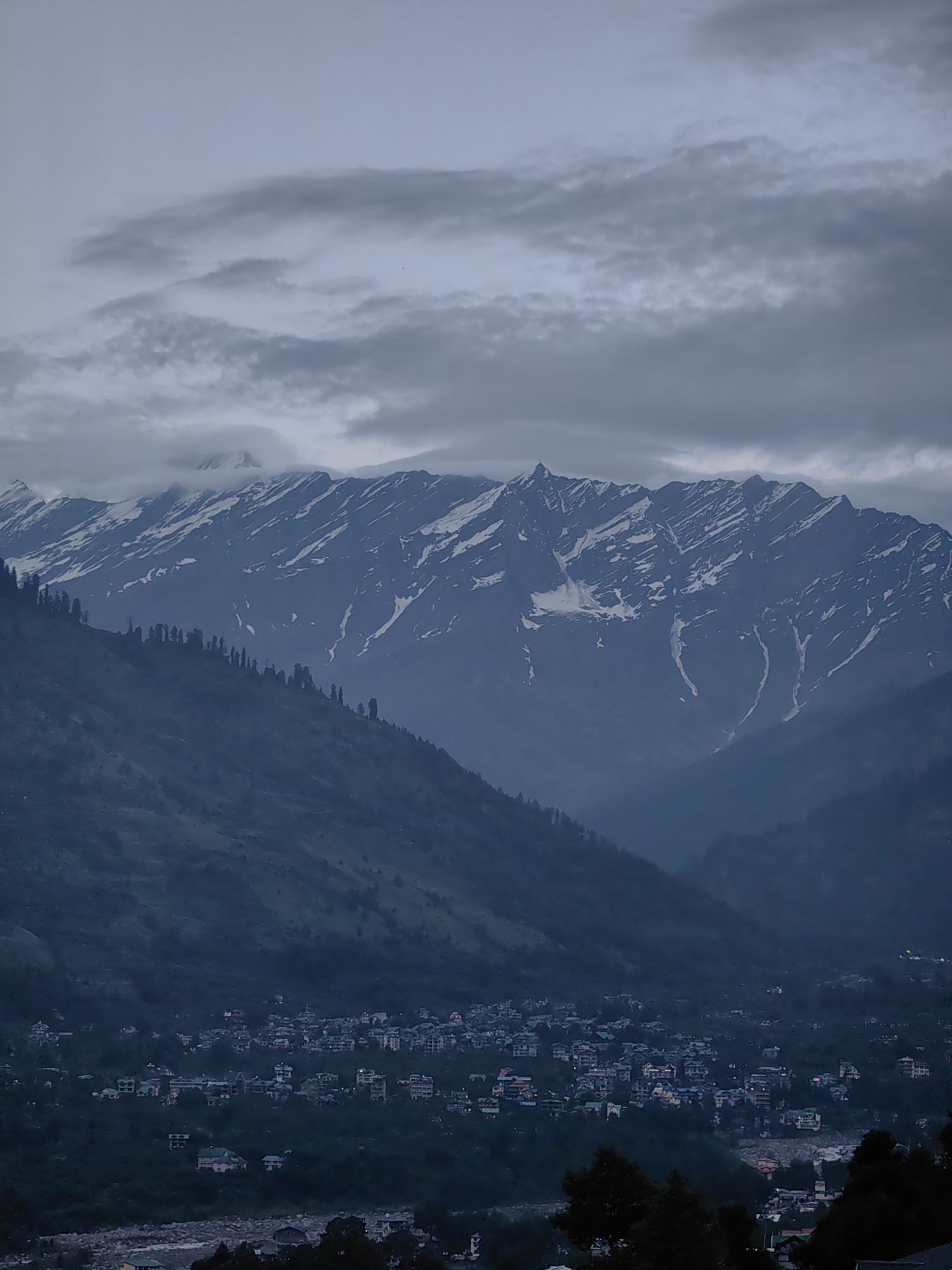 The image captures a panoramic view of the mountainous terrain surrounding Vashisht, India, under a heavily overcast sky. In the foreground, a dense collection of buildings, characteristic of a town or village, is nestled on a verdant mountainside. The architecture appears to be a mix of multi-story structures with visible windows and balconies, suggesting residential or commercial use. Some buildings are illuminated, indicating human presence and activity within the settlement.
Further up the slope, a line of tall, slender trees stands silhouetted against the darker landscape. Beyond this, the imposing presence of a mountain range dominates the upper portion of the image. The peaks are rugged and appear to be snow-capped, with streaks of white snow contrasting against the dark rock. Wisps of clouds cling to the mountainsides and the sky is filled with thick, grey cloud cover, creating a muted and somber atmosphere. The overall impression is one of a remote, high-altitude settlement overshadowed by the grandeur and ruggedness of the natural environment.