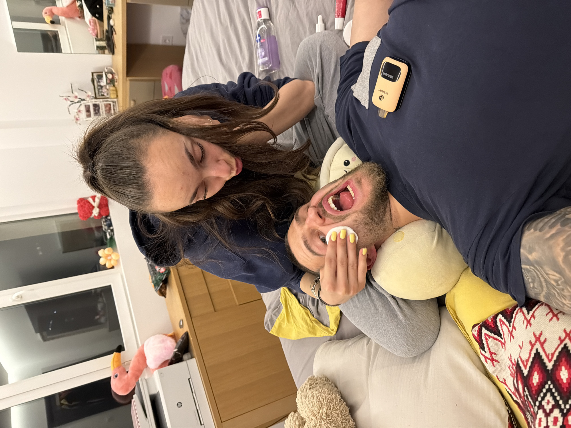 A man and woman are lying on a bed in what appears to be a bedroom. The woman is positioned above the man, leaning down with her face close to his. She has long, brown hair and is wearing a dark blue hoodie. The man, who has a beard, is lying with his mouth open, and the woman is holding a cotton pad near his face. He is wearing a grey shirt and is lying on a yellow pillow. A device that says “vdebeer” is clipped to his shirt, along with gray sweatpants. A blanket, lotion, and other products are on the bed with them. The bed has white linens. The camera is positioned at an angle, looking down on the scene. A cabinet with a pink flamingo and other items is in the background. It is not possible to tell where the image was taken.