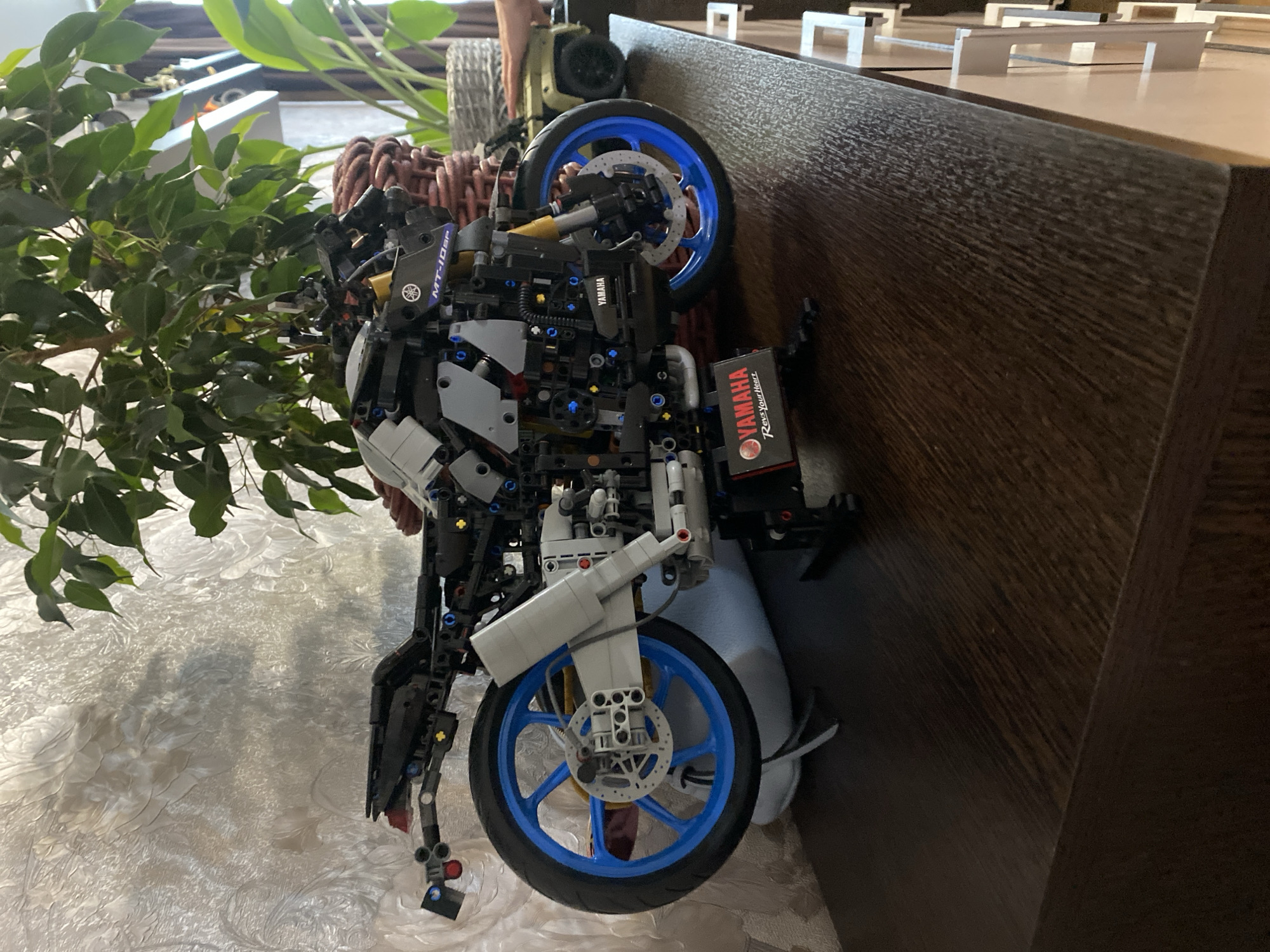 A detailed Lego Technic model of a Yamaha MT-10 SP motorcycle is displayed on a dark wooden surface. The motorcycle is black and gray with bright blue wheels. A Yamaha logo and "Rev Your Heart" are visible on a display plaque in front of the motorcycle. Behind the motorcycle, a large leafy green plant in a wicker basket is partially visible, as is a textured, light-colored wall. To the right of the plant, a small green toy vehicle and a hand are visible. The overall scene suggests an indoor display or collection. The location of "Берёзовая Роща, Russia" is not evident from the image itself.