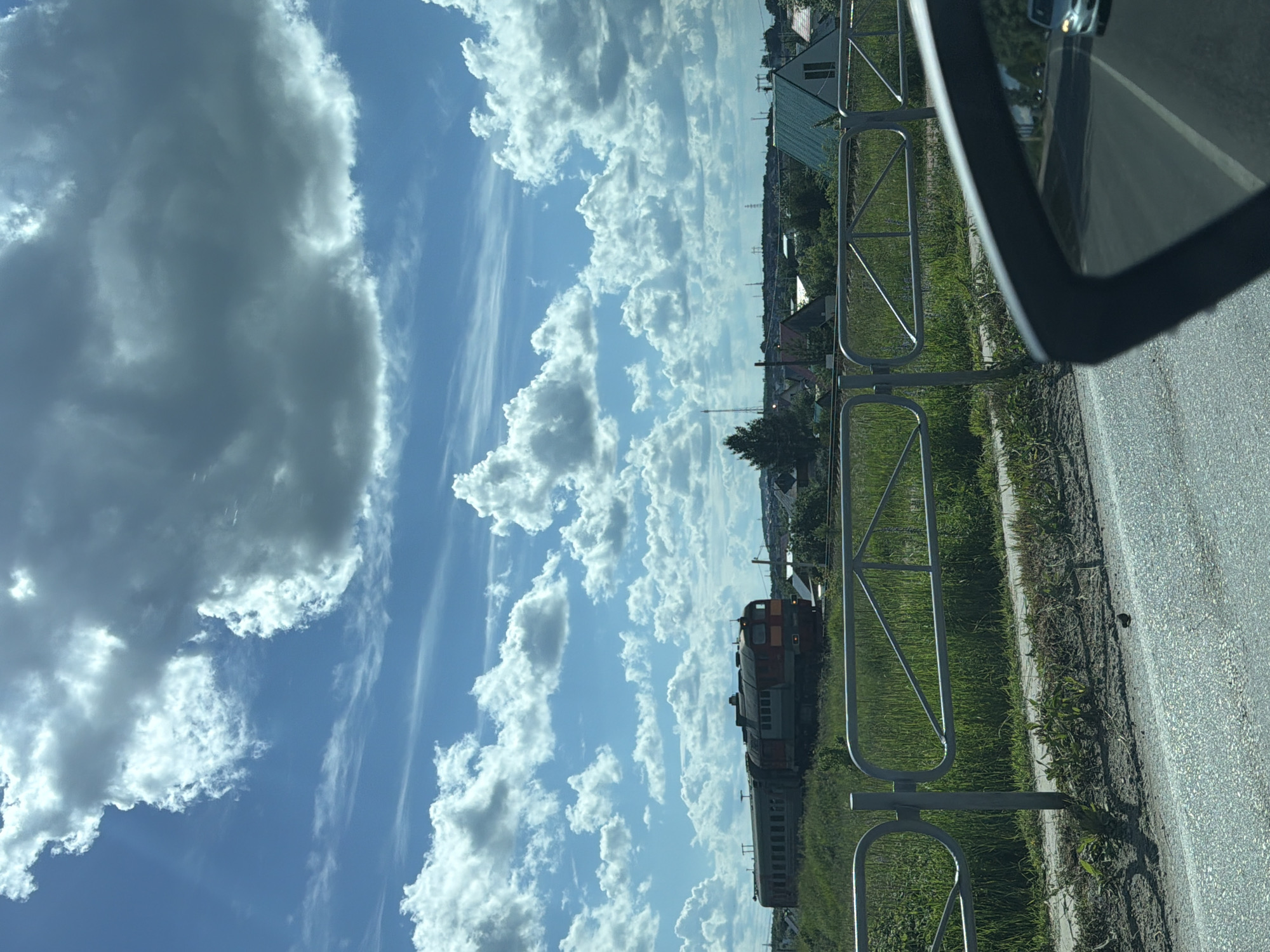 The image captures a view of a suburban or rural area, possibly on the outskirts of Chaikovsky, Russia, on a bright, cloudy day. A train, predominantly red and dark gray, is visible on tracks to the left of the frame, moving horizontally across the landscape. The train consists of several passenger cars. In the background, a cluster of residential buildings with various roof colors, including green and dark gray, can be seen, interspersed with trees and vegetation. A metal fence runs horizontally in the mid-ground, separating the foreground from the area with the train and buildings. The foreground is dominated by a paved road, with a car's side mirror reflecting a car in the distance. The sky is a vivid blue, filled with large, puffy white and gray cumulus clouds, suggesting dynamic weather conditions. The overall scene depicts a typical day in a location with both infrastructure and natural elements.