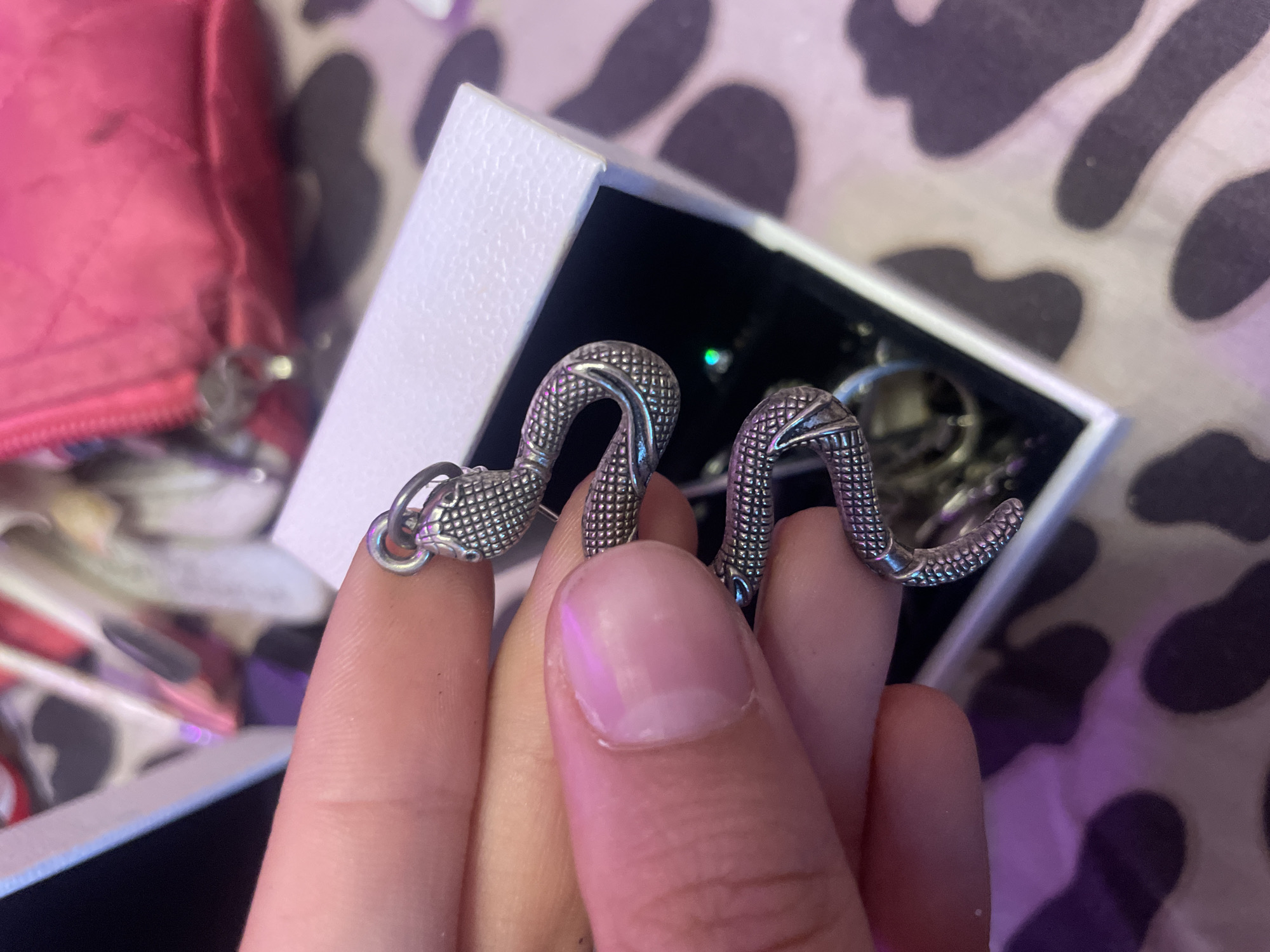 The image shows a person's hand holding a silver snake charm. The charm is detailed with scales and a textured pattern, with two snake heads visible. The charm is being held over a white jewelry box that is open, revealing other jewelry inside. In the background, a pink quilted bag is visible, and the surface below the box has a dark and light spotted pattern. The lighting in the image is somewhat dim with a purple and pink hue. The location is not Ladozhskoe Ozero, Russia; this image depicts an indoor setting with personal items.