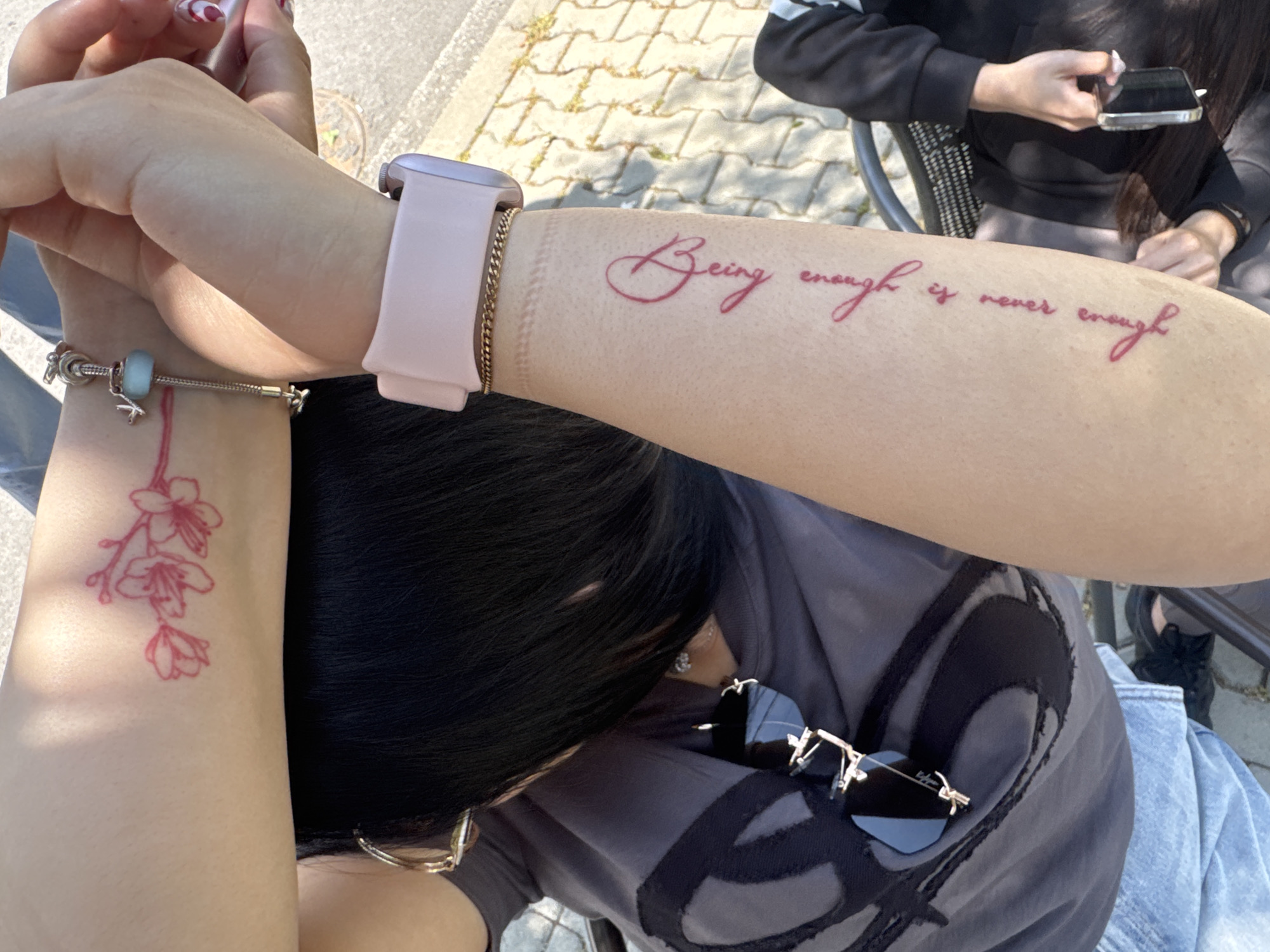 A medium shot shows a person seated outdoors with their arms crossed, displaying tattoos. The right forearm features a tattoo in cursive script that reads "Being enough is never enough", while the left wrist has a tattoo of cherry blossoms hanging downward. The person is wearing a pale pink Apple Watch on the right wrist and a silver charm bracelet on the left. They are wearing a dark grey t-shirt with a black design, and sunglasses are clipped to the t-shirt. The person has short black hair. In the background, another person is seated and using a cell phone. The environment appears to be an outdoor seating area with a light-colored paved surface visible in the background.
