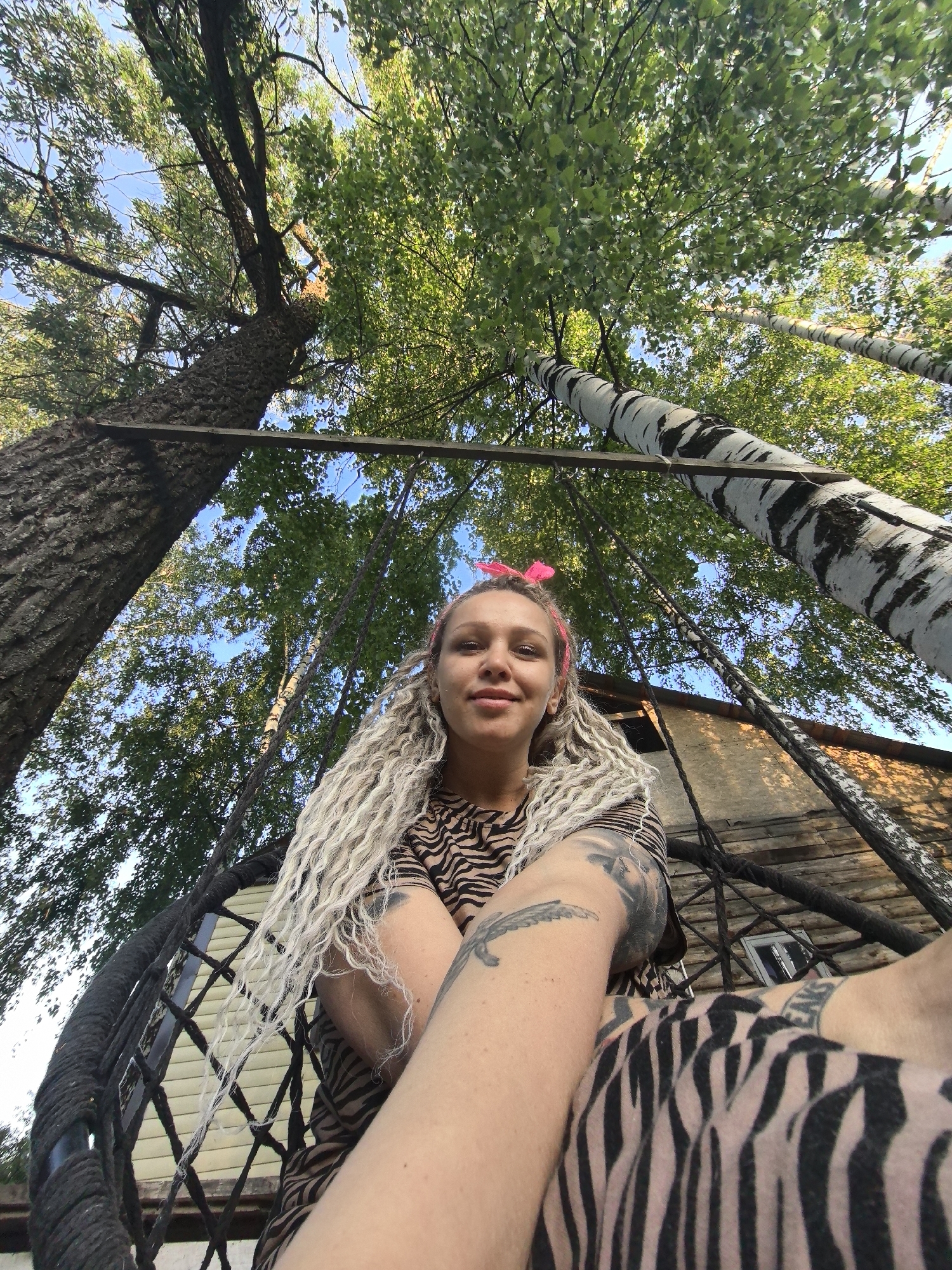 A low-angle shot depicts a woman with long, blonde dreadlocks and a pink bow headband sitting in what appears to be a swing or hanging chair. She is wearing a brown and black zebra-print top. Her arms are adorned with tattoos. The camera is positioned below, looking up towards her face.
The backdrop consists of trees. A large, bark-covered tree on the left, while two birch trees with their characteristic white and black markings are visible on the right. The foliage forms a canopy overhead, with glimpses of a light blue sky peeking through. The structural support, possibly for the swing, is visible between the trees. A wooden structure can be seen in the background, behind the birch trees.
The location is Noviy, Russia.
