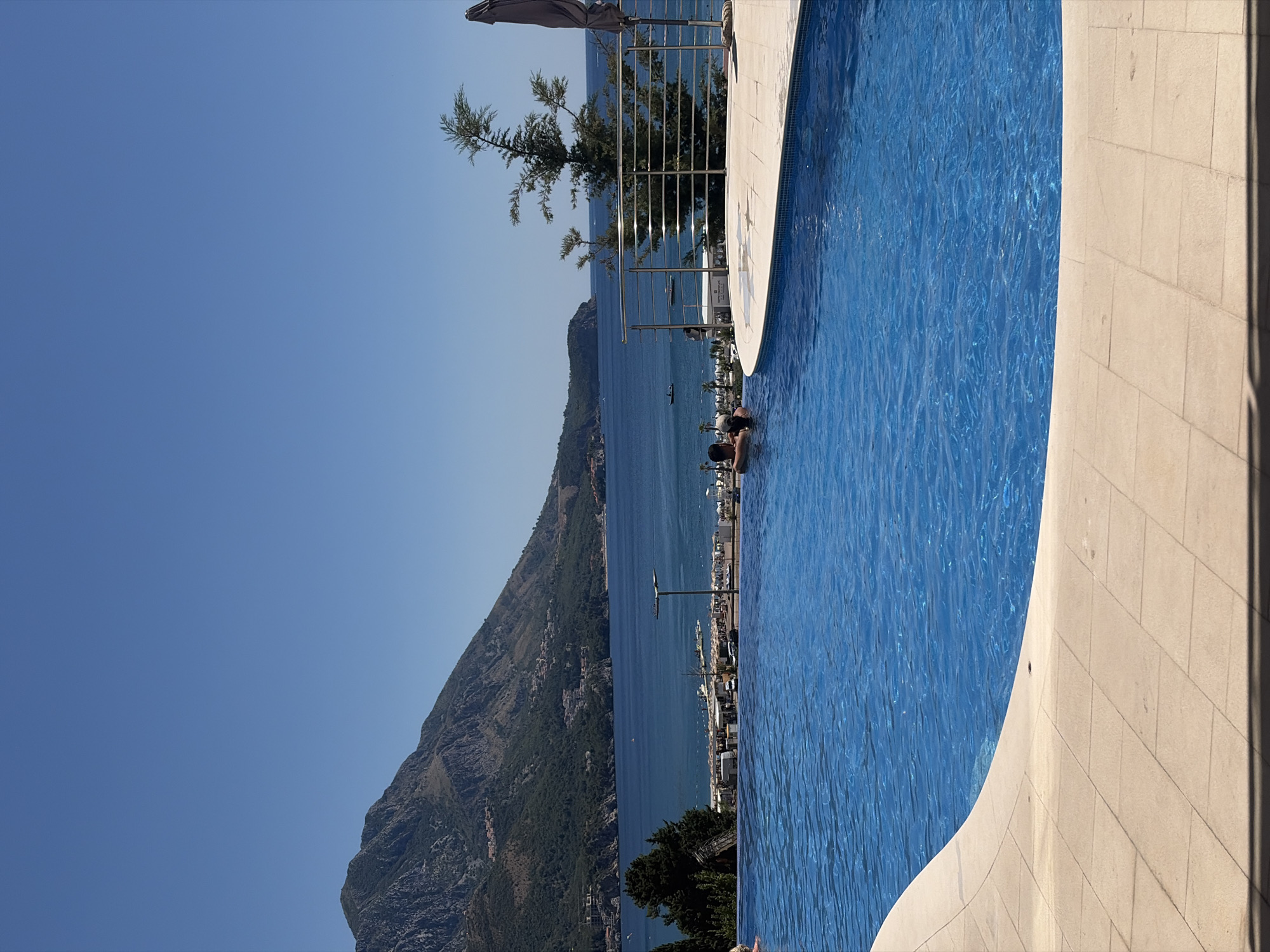 An infinity pool overlooks a bright blue sea and a mountainous coastline under a clear sky. Two people are in the pool, partially submerged, embracing. Beyond the pool, a beach with rows of umbrellas and lounge chairs is visible, with a few boats on the water. The background features a lush green mountain range dotted with buildings, characteristic of the coastal towns in Montenegro. A tree and a closed umbrella are near the pool's edge. The poolside is paved with beige tiles.