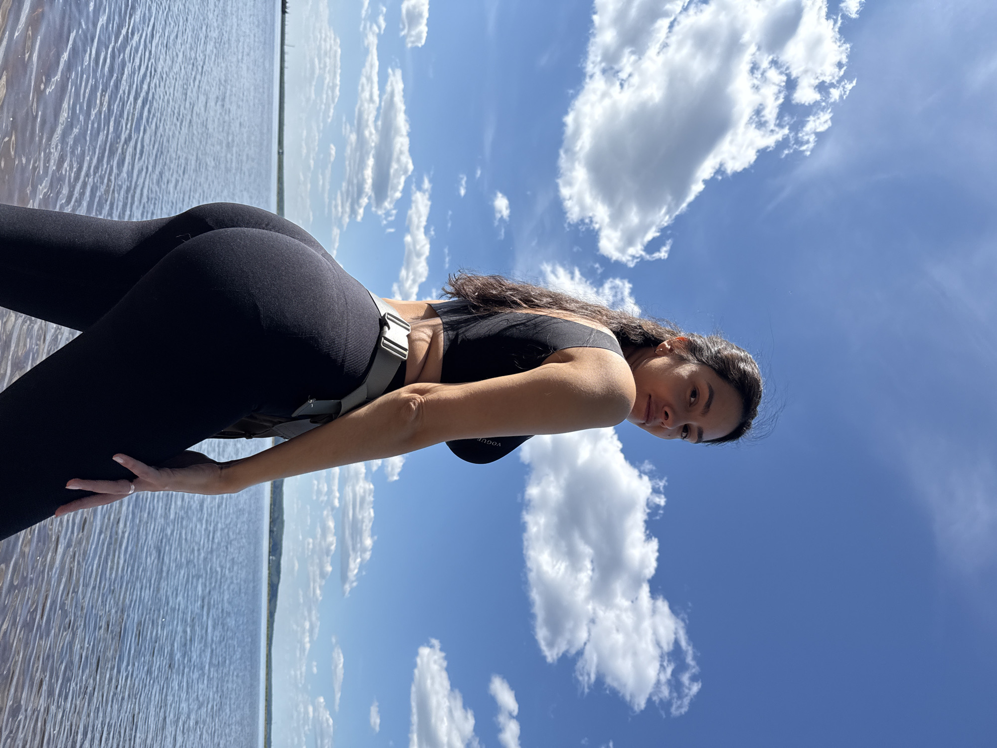 A female subject with dark, wavy hair is depicted from the waist up, standing with her back partially turned to the camera, twisting at the torso to look over her right shoulder directly into the lens. She has a neutral expression and wears a black athletic crop top or sports bra with faint "VOGUE" branding on the front, and black high-waisted leggings. A gray webbed belt with a prominent metallic quick-release buckle is visible around her waist. Her left hand, adorned with a ring on the fourth finger, rests on her left thigh.
The setting is an outdoor, sunny environment with a clear blue sky sparsely populated by scattered white cumulus clouds. The lower half of the frame is dominated by a large body of water, exhibiting ripples on its surface. A low-lying landmass or distant shoreline is visible on the horizon across the water. On the far left of the distant horizon, indistinct structures, possibly industrial, are faintly discernible. The subject is positioned at the water's edge, consistent with a large reservoir or riverbank environment, such as those found near the city of Chaikovsky, Russia.