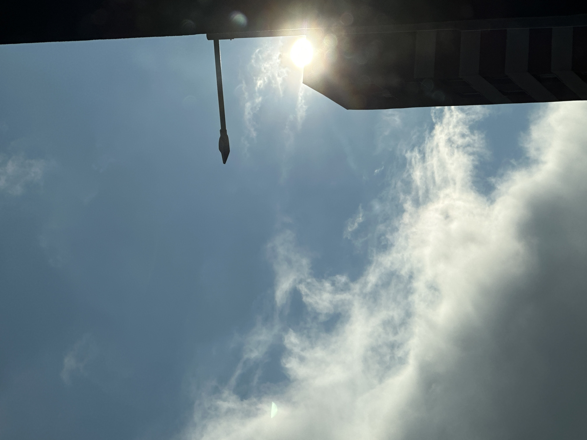 A low-angle shot of the sky in Sofia, Bulgaria. A long, thin, dark object with a pointed end hangs from a dark, shadowed structure at the top left of the frame. The sun shines brightly near the top center, partially obscured by the structure. The sky transitions from a light blue at the left to a cloudier area on the right. Wispy white clouds are scattered across the blue sky, with a denser cloud mass visible on the right side. Dark structures are visible at the top right of the frame.