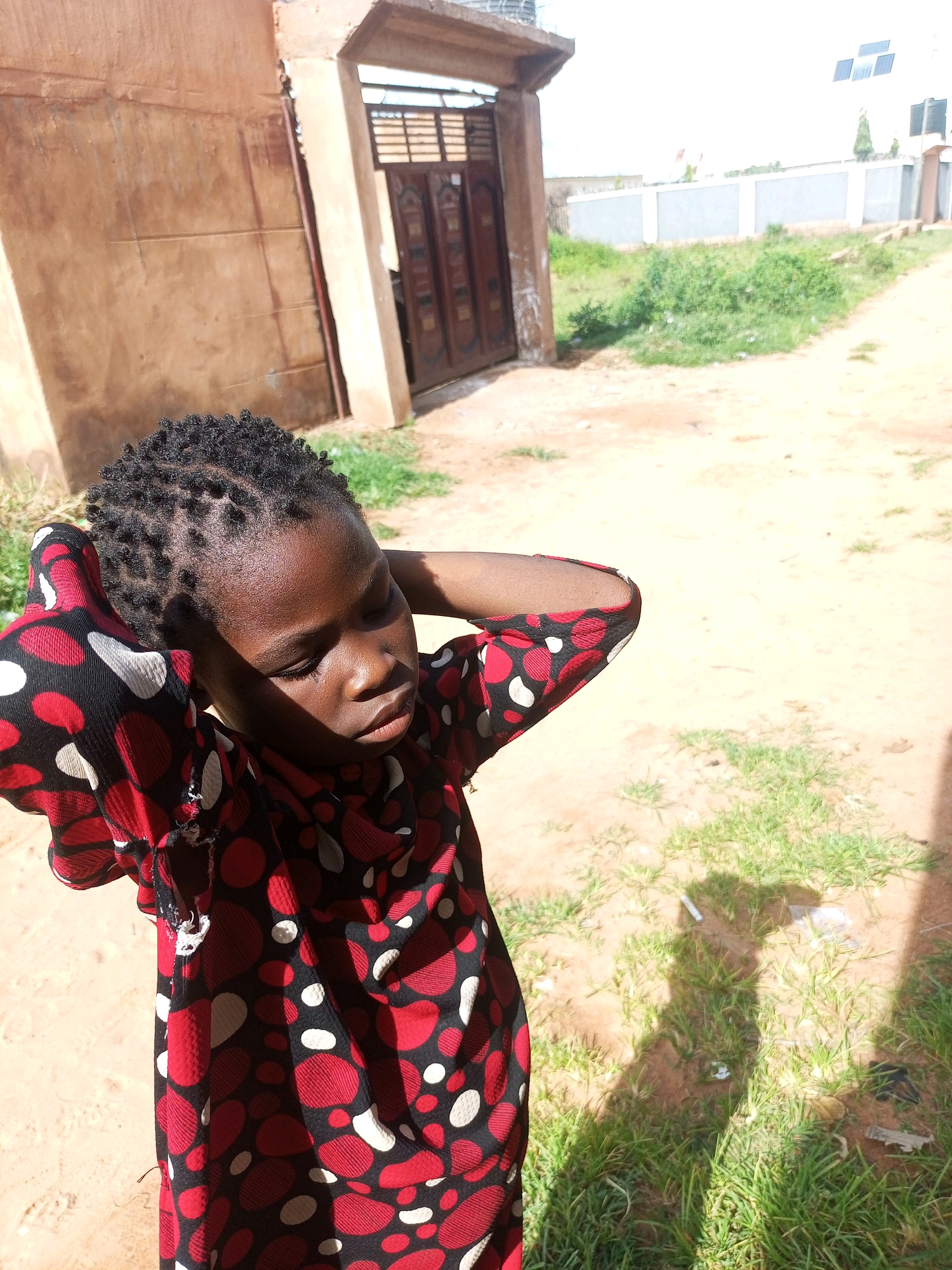 A young African girl stands outdoors, her arms behind her head, with her eyes closed and a peaceful expression. She has her hair braided into small twists. She is wearing a red, black, and white polka-dotted dress. The background features a dirt path with patches of grass. To the left is a simple, light brown building with a dark brown wooden door. To the right, further in the distance, is a modern white building with solar panels on the roof and a grey fence. The lighting suggests it is daytime, with sunlight casting shadows on the ground. The scene appears to be in a rural or semi-urban area of Tempure, Nigeria.