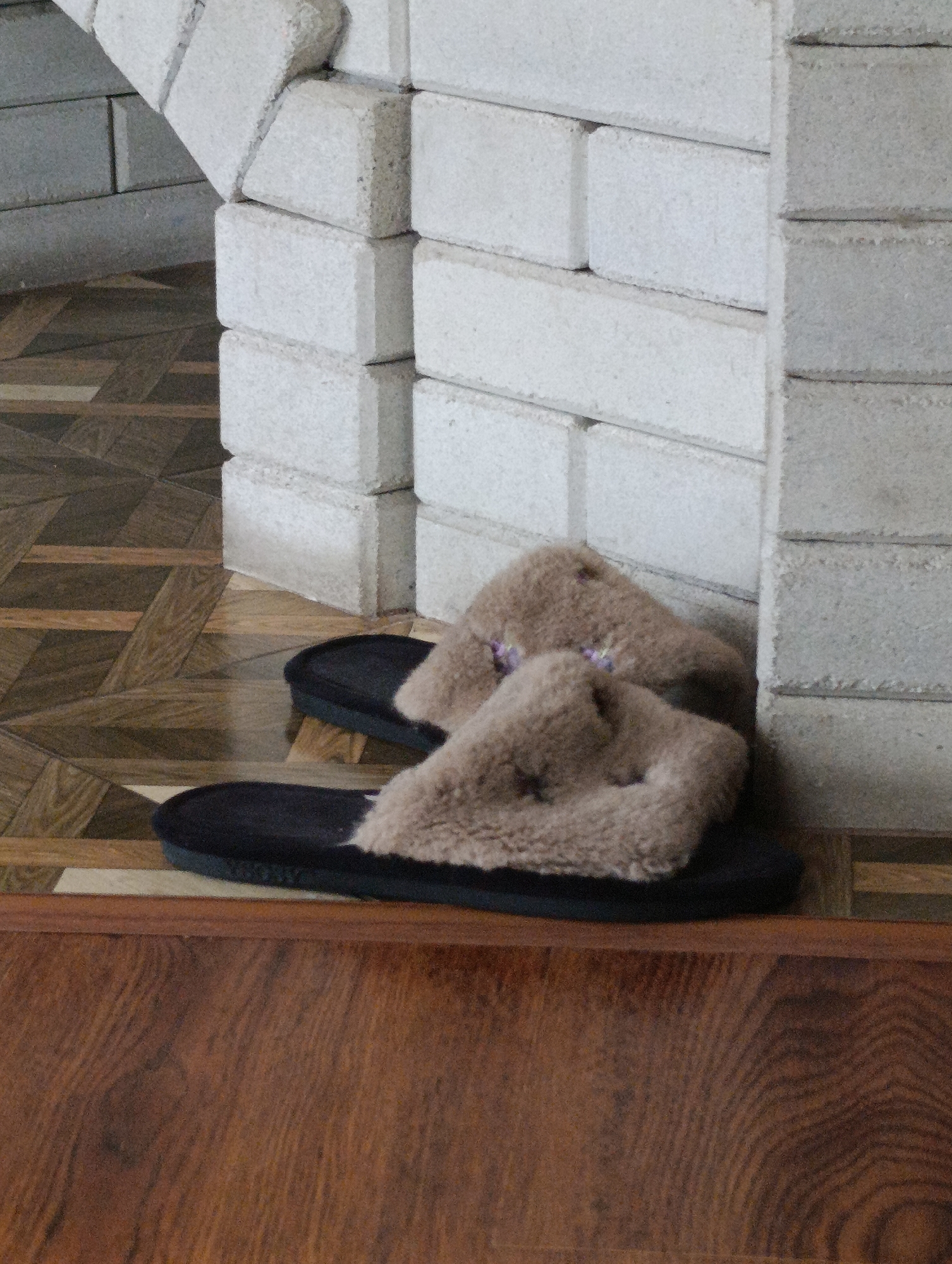A pair of fluffy, tan-colored slippers with black soles rests on a wood-paneled floor. The slippers are positioned near a wall constructed of light grey bricks, which forms an archway on the left side of the frame. The slippers are the primary focus of the image. The background consists of the brick wall and the patterned floor. There are no people or any other discernible objects in the immediate vicinity of the slippers. The scene appears to be indoors, possibly in a residential setting.