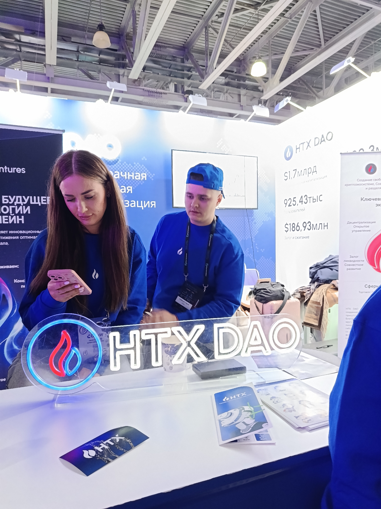 Here is a detailed description of the image:
**Setting:** The image appears to be taken at a conference or trade show in an indoor space. The location is not specified. There is a backdrop or display behind the table with marketing materials in Russian.
**People:** Two individuals are in the frame. One is a young woman with long hair, wearing a blue sweatshirt. She is looking at a pink smartphone she holds in her hand. The second person is a young man with a blue baseball cap and a blue sweatshirt. He has an identification badge around his neck and is looking towards the right side of the frame.
**Objects:** Several objects are visible on the table. A neon sign displays the text "HTX DAO" with a graphic logo. There are brochures and promotional materials with the HTX logo and additional printed materials on the table. Behind them, there are suitcases and bags on the floor behind the table.
**Actions:** The woman is interacting with her phone, while the man is looking in a different direction, possibly at someone off-camera. They appear to be working at a booth.
**Environment:** The environment suggests an indoor setting with artificial lighting, possibly at a convention center. Structural elements like metal beams are visible on the ceiling. The overall ambiance is that of a professional or business event.
**Subject:** The primary subject is the HTX DAO marketing presence at the event, including the promotional materials and the booth staff.