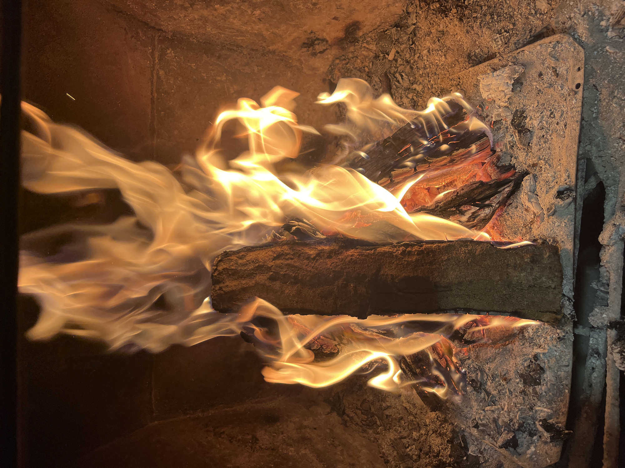 The image shows a fire burning inside what appears to be a wood-burning stove or fireplace. There are logs actively burning with visible flames. Ash and remnants of burnt wood are present in the bottom of the fireplace. The interior walls of the fireplace are rough and textured. The image does not show any people, nor does it indicate that it was taken in Varshilo, Bulgaria.