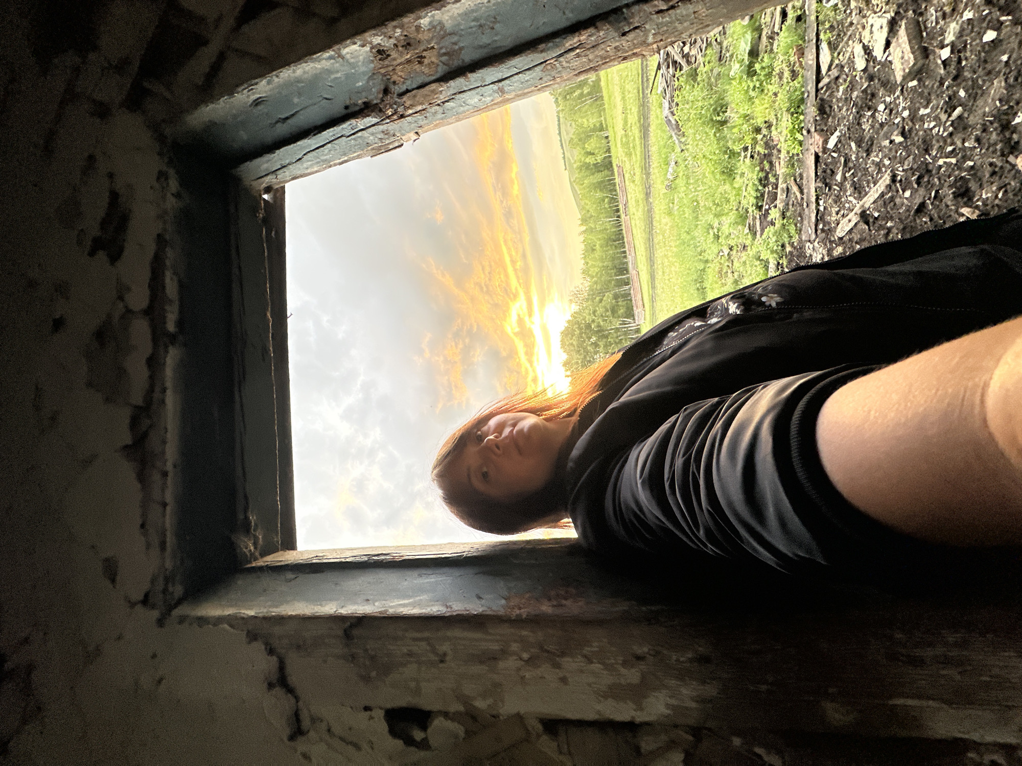 A person with shoulder-length dark hair and orange highlights is leaning against a wall inside a dilapidated structure, looking towards the camera. They are wearing a black jacket. The person is holding their arm out, as if taking a selfie. Through an opening in the structure, a rural landscape with green fields, trees, and a colorful sunset sky is visible. The opening is framed by rough, weathered walls and a rusty metal frame. The ground inside the structure is littered with debris.