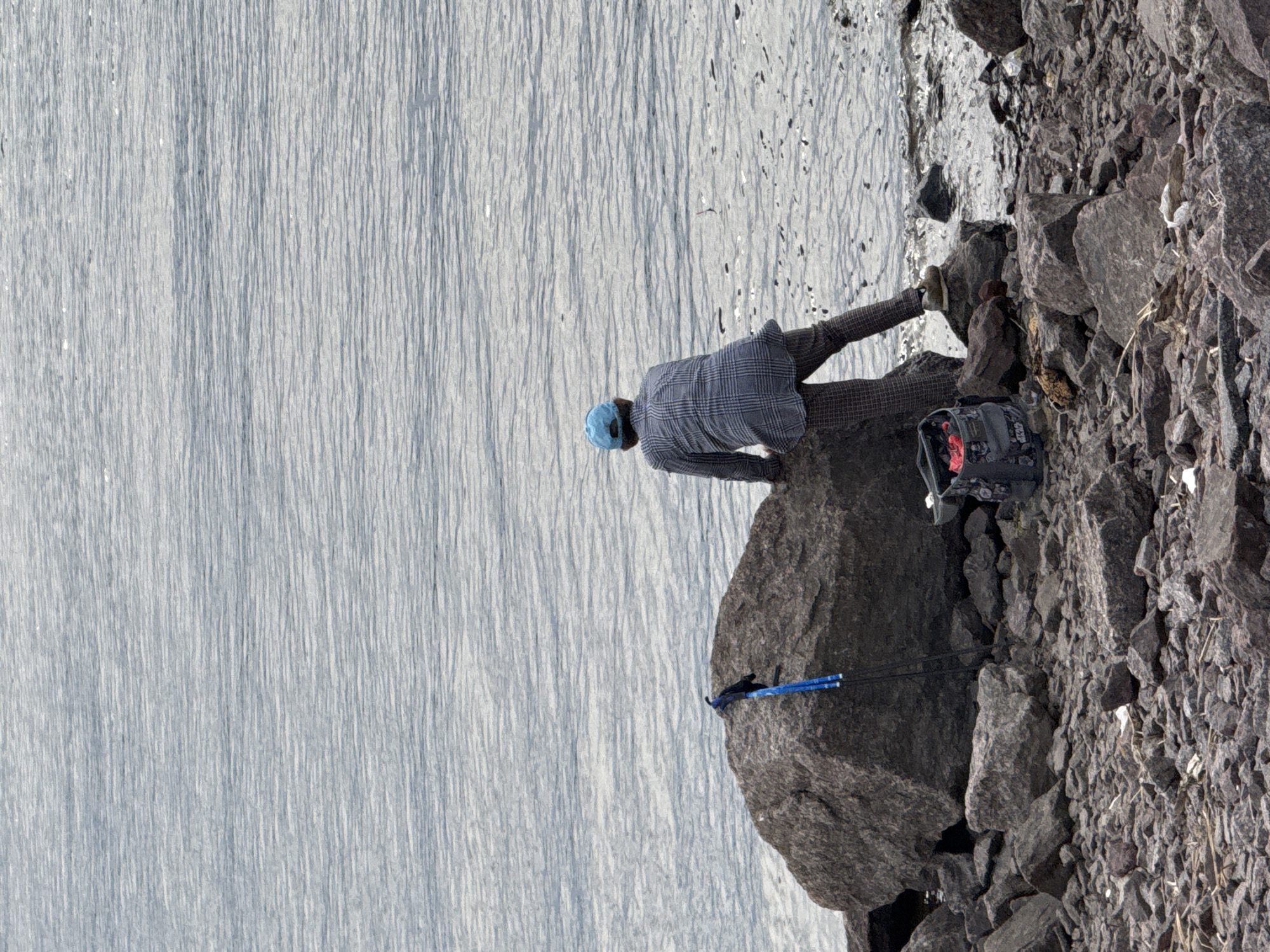 A person in a blue cap and plaid shirt stands on a rocky shoreline next to calm water. A backpack and hiking poles rest on the rocks. The location appears to be a natural environment, but it is within the city of Saint Petersburg, Russia, based on the presence of granite and the proximity to a body of water, which is characteristic of the Neva River delta.