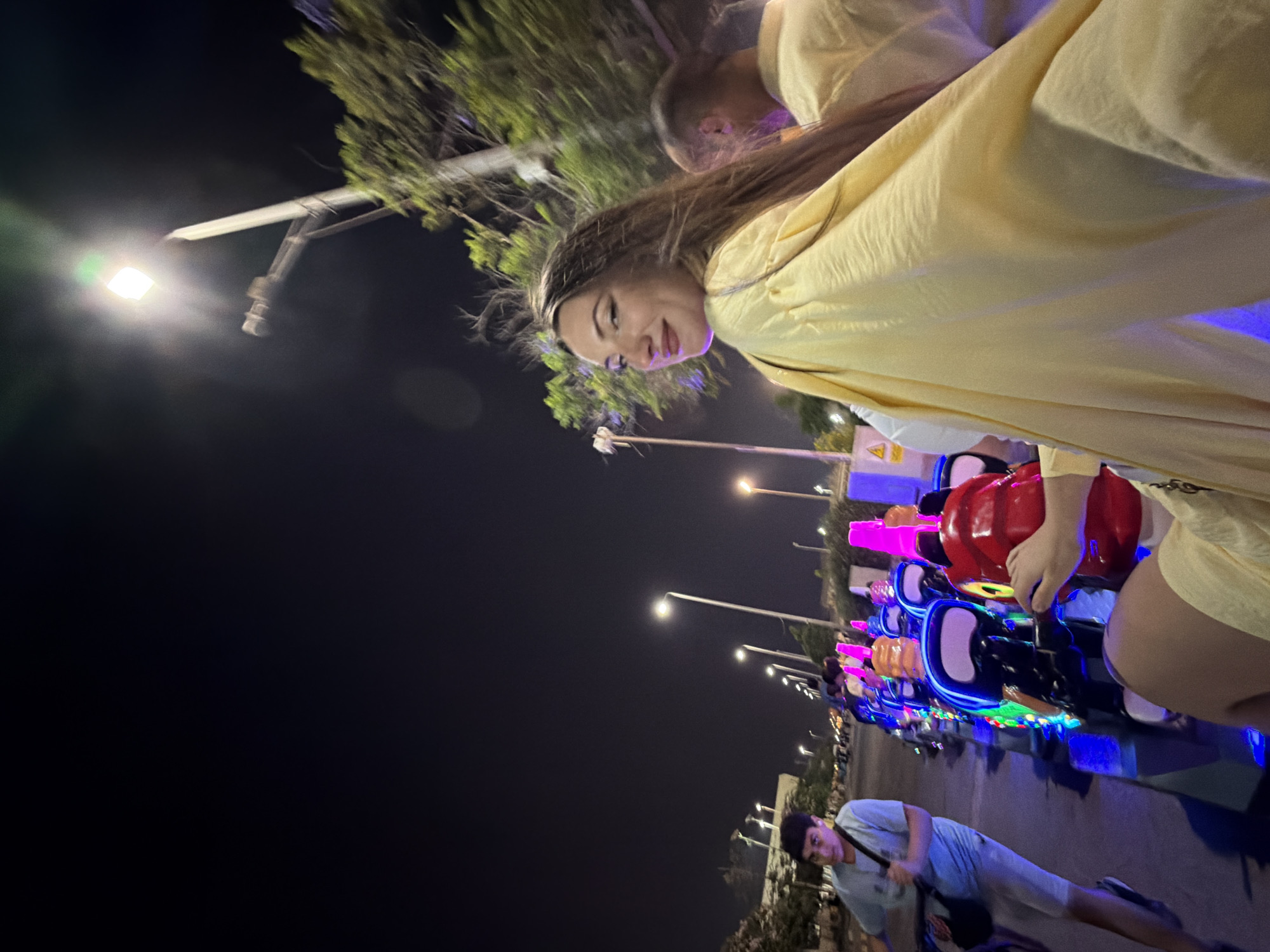 At night in the city of 6th of October, Egypt, a group of people ride in a line of illuminated vehicles. A young man in a light blue t-shirt and shorts is in the foreground on the left, looking forward. To his right, a woman with long brown hair wears a light yellow garment and smiles at the camera. She is seated in one of the vehicles, which are brightly lit with blue and pink neon lights. Another person in a light yellow top is visible behind her. The vehicles appear to be a type of recreational ride, possibly small go-karts or similar novelty vehicles. Streetlights illuminate the scene, and trees are visible in the background. The overall atmosphere suggests a nighttime entertainment venue or public space.