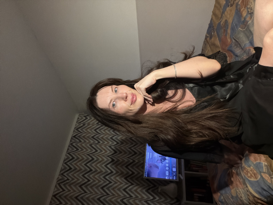 A woman with long brown hair rests on a bed with a patterned comforter. She wears a black silky robe and a silver bracelet. Her hand is gently placed near her face. In the background, a chevron-patterned wall and a TV are visible, sitting atop a shelf filled with books. The room is dimly lit.