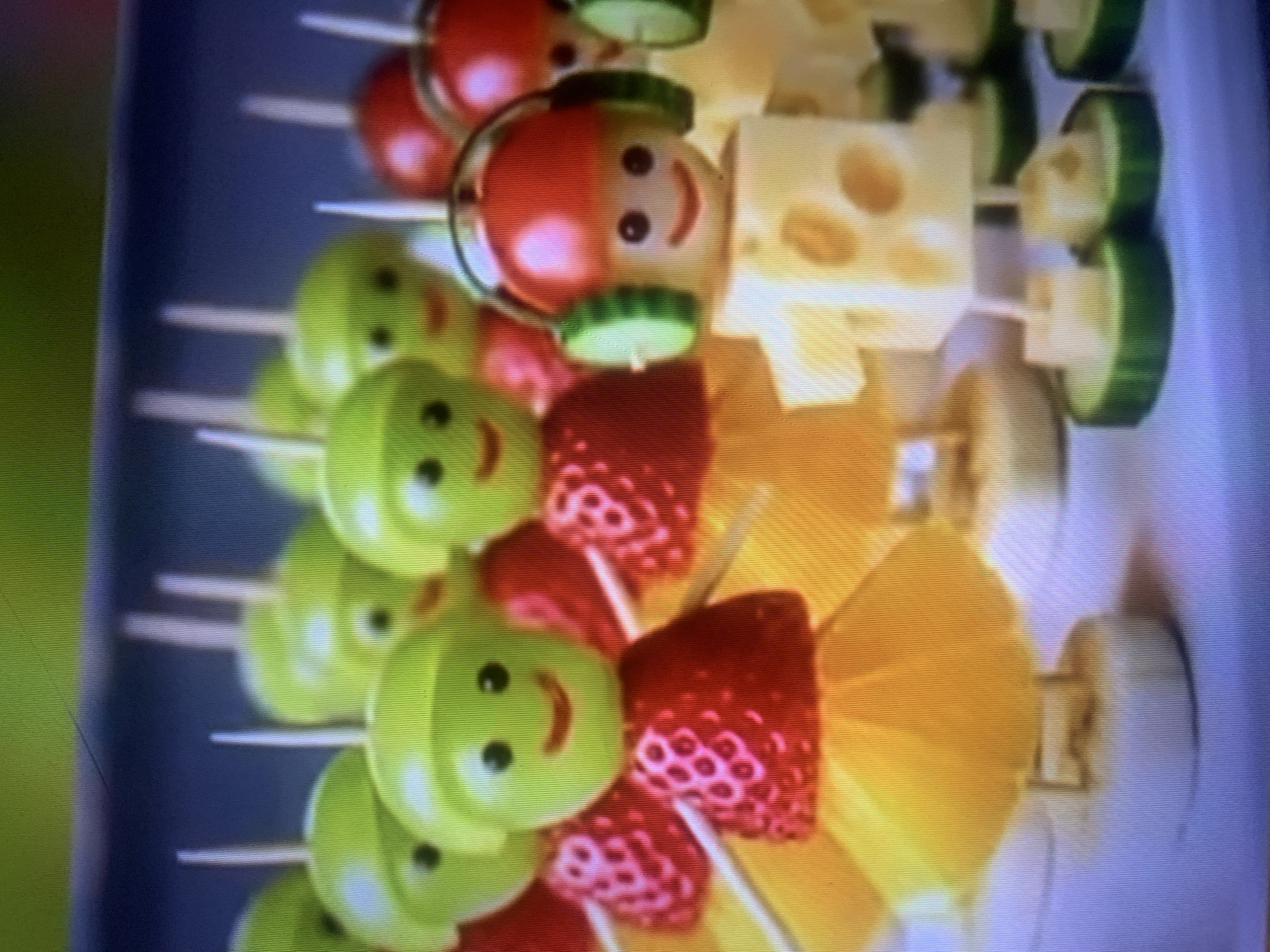 The image displays a close-up, high-angle shot of a collection of fruit and cheese skewers arranged on a surface. The skewers are crafted to resemble characters. The characters have heads made from green grapes or cherry tomatoes, with black edible dots for eyes and a red piece for a mouth. Their bodies are composed of strawberries, orange slices, and cheese cubes. Some characters are adorned with cucumber slices as headphones. The skewers are planted upright, with their bases resting on slices of banana and cucumber. The background is out of focus, showing a hint of a blue surface and possibly some green at the top. The scene depicts a prepared food arrangement, likely for a party or event. The location is not identifiable from the visual information provided, and there is no indication of the city of Almaty, Kazakhstan.