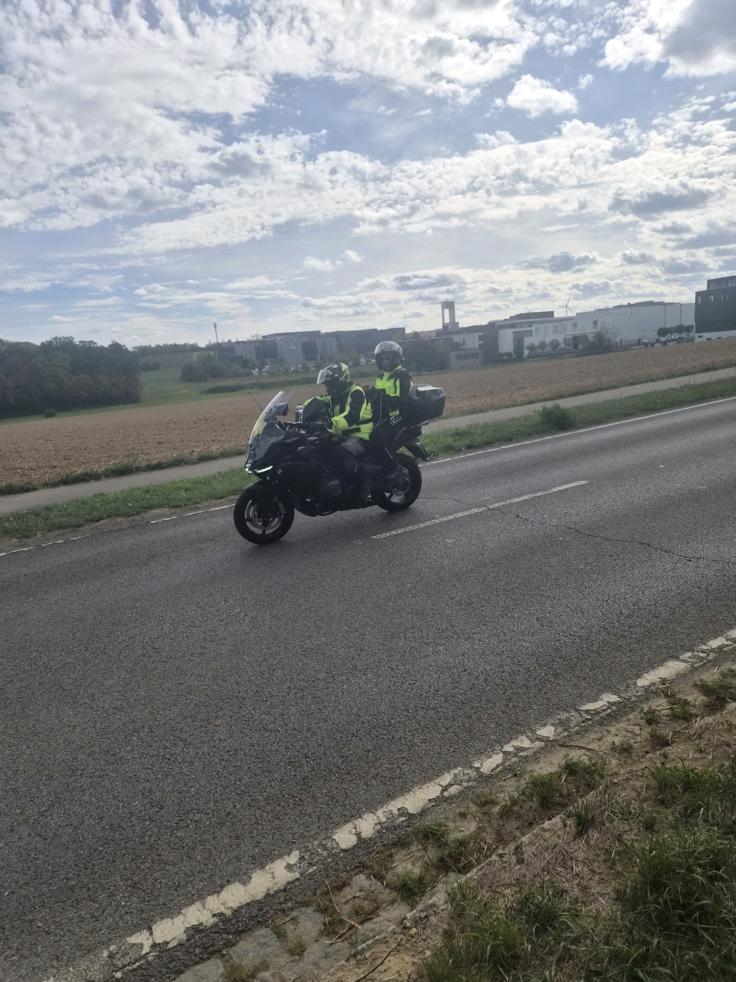 The image shows a black motorcycle with two riders wearing high-visibility vests and helmets, traveling on a paved road. The motorcycle has a top box and a windshield. The riders are seated one behind the other. In the background, there are fields, trees, and buildings that appear to be industrial or commercial structures. The sky is partly cloudy. The road has a white line marking. The setting is likely Leudelange, Luxembourg, given the context.