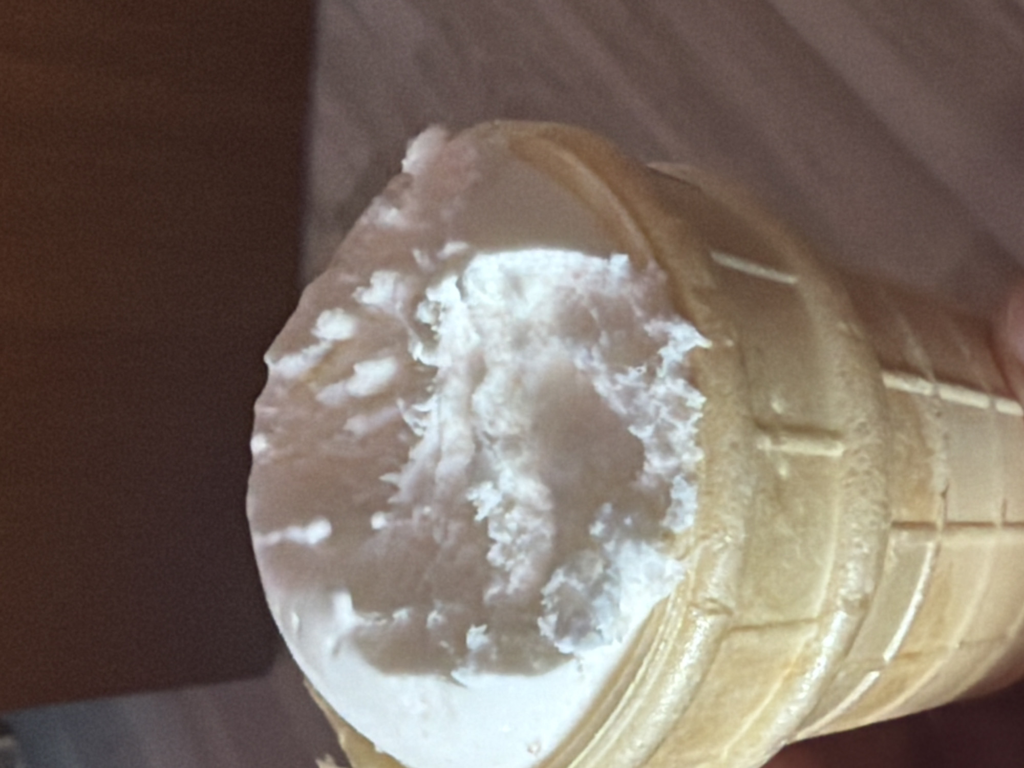 A close-up, eye-level shot shows a vanilla ice cream cone. The ice cream is a pale white, with a slightly rough texture and a few visible air pockets. It is partially melted, with some soft peaks and drips. The waffle cone is a light golden brown and has a textured grid pattern. The background is out of focus and appears to be a dark, brown surface. The image is taken indoors, likely in a casual setting.
