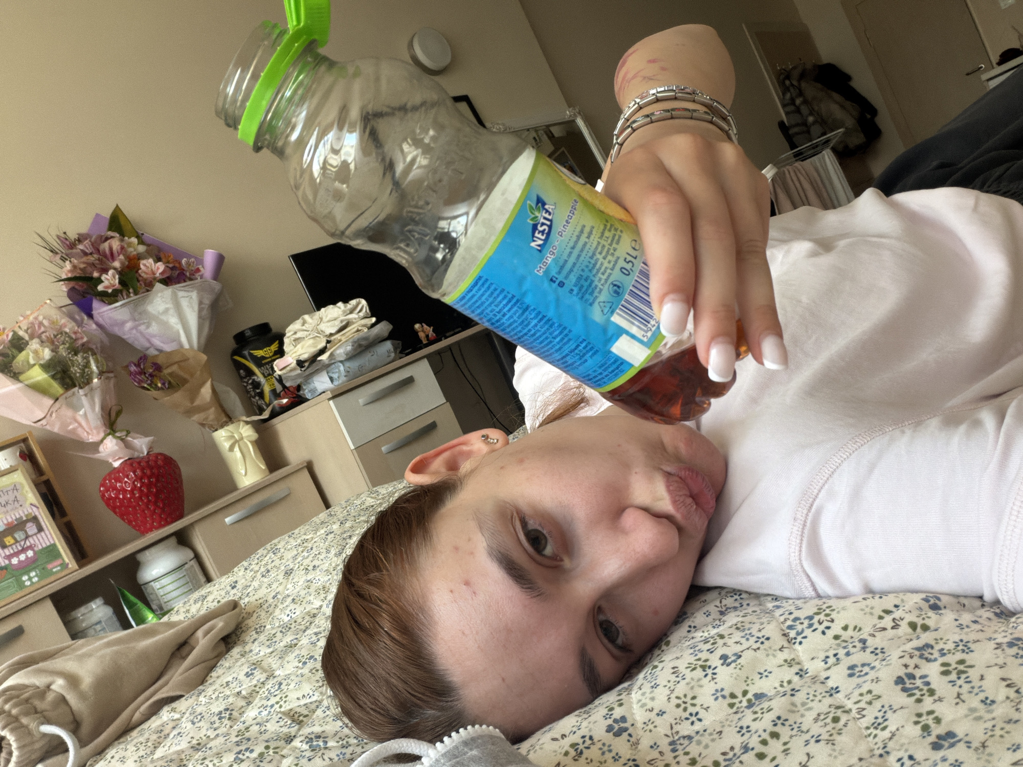 A young woman with reddish-brown hair and a slight pout lies on her back on a bed covered with a light floral-patterned duvet. She is wearing a light-colored top and holding an almost empty plastic bottle of Nestea Mango-Pineapple juice upside down over her face, as if trying to extract the last drops. Her hand displays a French tip manicure and several silver-toned bracelets.
The indoor setting appears to be a bedroom. In the background, on a dresser or shelf, multiple bouquets of cut flowers are visible, along with a prominent red strawberry-shaped decoration, a cream-colored vase, and various other bottles and containers. A framed picture with indistinct Cyrillic text and a drawing of buildings is also on the shelf. A wardrobe or cabinet and what appears to be a television or monitor are partially visible in the further background.
Visible text on the bottle label includes "NESTEA", "Mango-Pineapple", "0.5LE", and "NESTLÉ", along with a barcode and smaller text indicating ingredients and nutritional information in multiple languages. The scene suggests a relaxed moment at home, potentially in Sofia, Bulgaria.