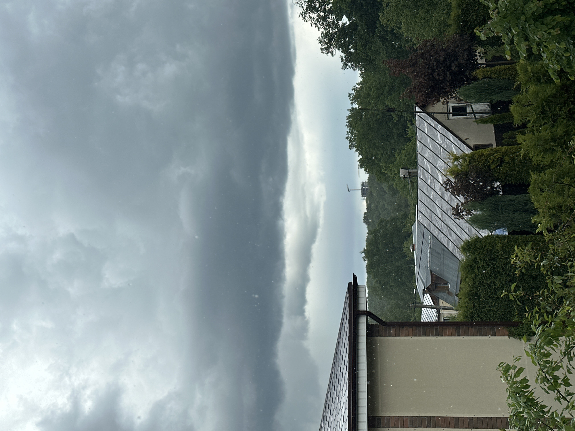 The image shows a stormy sky above a residential area in Nizhny Novgorod, Russia. A dark, heavy band of clouds dominates the left side of the frame, contrasting with a lighter, brighter patch of sky in the center. Rain or snow is falling, visible as small white specks throughout the scene. Trees are visible on the right, bordering the houses. Two houses with metal roofs are partially visible. A construction crane can be seen in the distance.