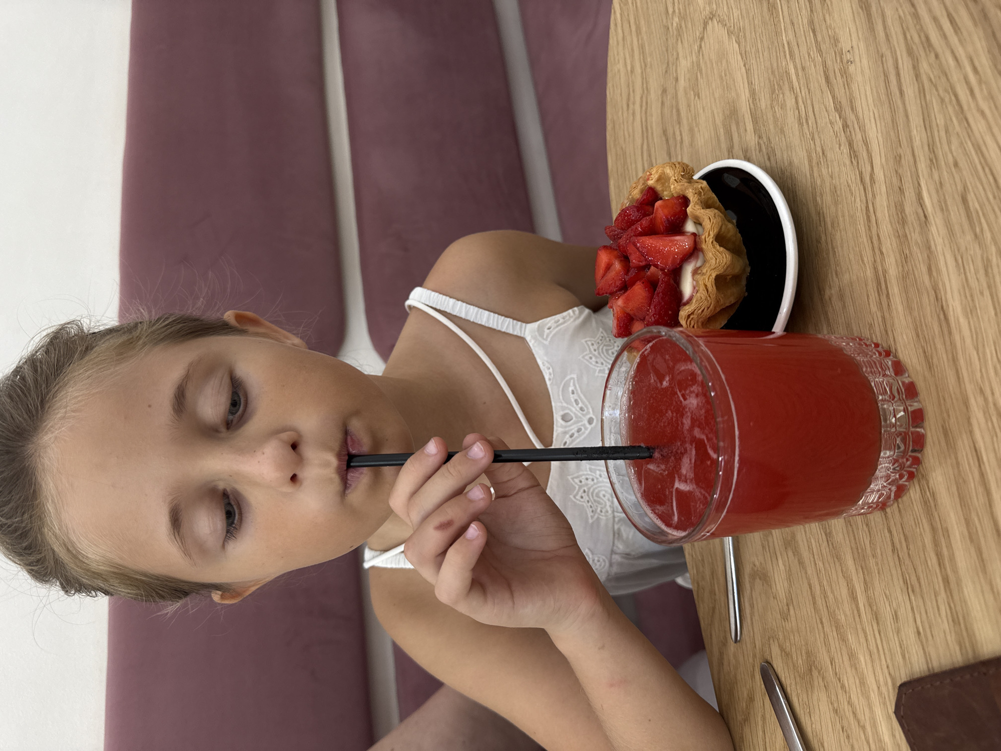 A young girl with her hair in a bun is drinking a red beverage through a black straw from a fluted glass on a wooden table. She is wearing a white dress with thin straps. To her right, on a black saucer, is a small pastry tart topped with fresh strawberries and a creamy filling. A silver fork is visible to the left of the glass. The background features a plush, deep pink seating area. The scene appears to be indoors, possibly in a cafe or restaurant.