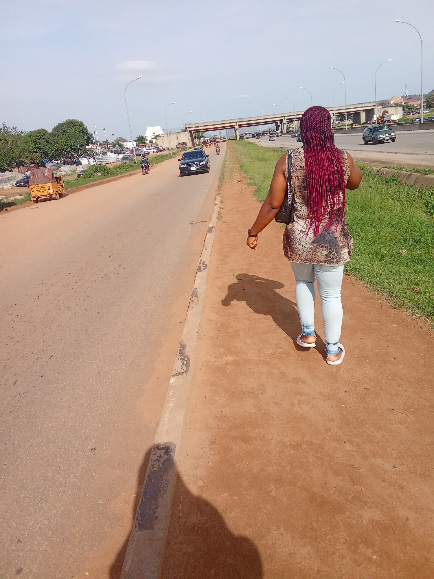 A woman with long, burgundy braids walks on a dirt path next to a paved road in Kagini, Nigeria. She is wearing a patterned sleeveless top and light blue jeans. A dark purse is slung over her left shoulder. The road has multiple lanes of traffic with several cars and a motorcycle visible. In the background, an overpass spans the road, and buildings and trees are scattered in the distance. The sky is clear and blue. To the left of the road, there is a yellow three-wheeled vehicle. The overall environment appears to be an urban or semi-urban area with ongoing infrastructure development, as evidenced by construction materials on the left side of the road.