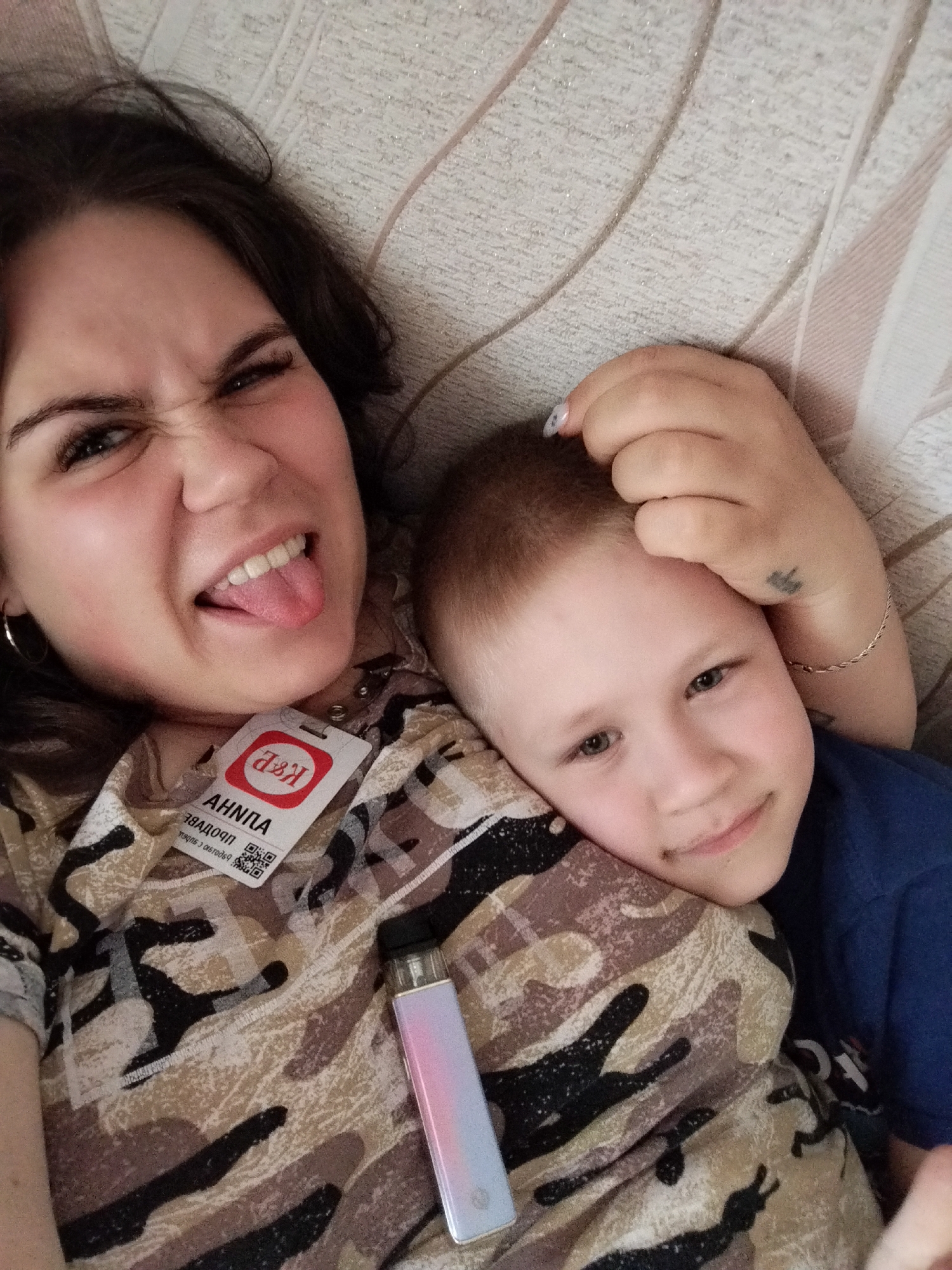 Two individuals, an adult female and a child, are lying down together. The female, positioned on the left, is making a playful face with her tongue sticking out. She is wearing a camouflage-patterned shirt with a lanyard and ID badge around her neck. The child, on the right, is resting his head on the female's shoulder, looking towards the camera with a gentle smile. A vape device is placed on the female's shirt. The background appears to be a textured, light-colored surface, possibly a sofa or bed. The image captures a moment of casual interaction and affection between the two. The location is the city of Chaikovsky, Russia.