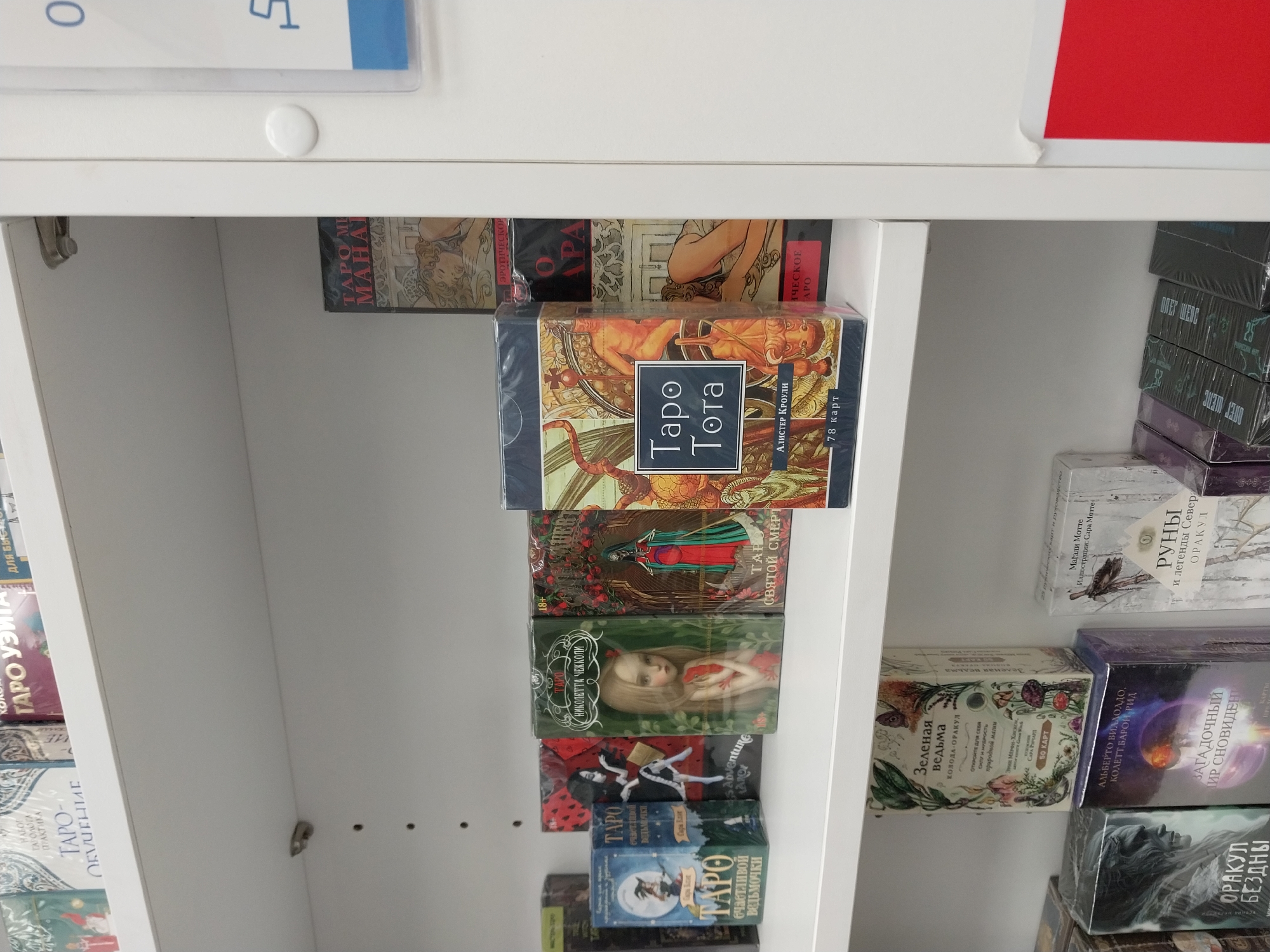 The image shows a shelf in a store, possibly in Sochi, Russia, based on the Cyrillic text on the various tarot card decks and books. The shelf is white and contains multiple decks of tarot cards and related books. The decks vary in design and title, and they are stacked one behind the other on the shelf. Some of the titles are visible, including "Taro Tota", "Green Witch", "Runes and Legends of the North", "Oracle of the Abyss", "Oracle of Dreams". Some decks are shrink-wrapped.