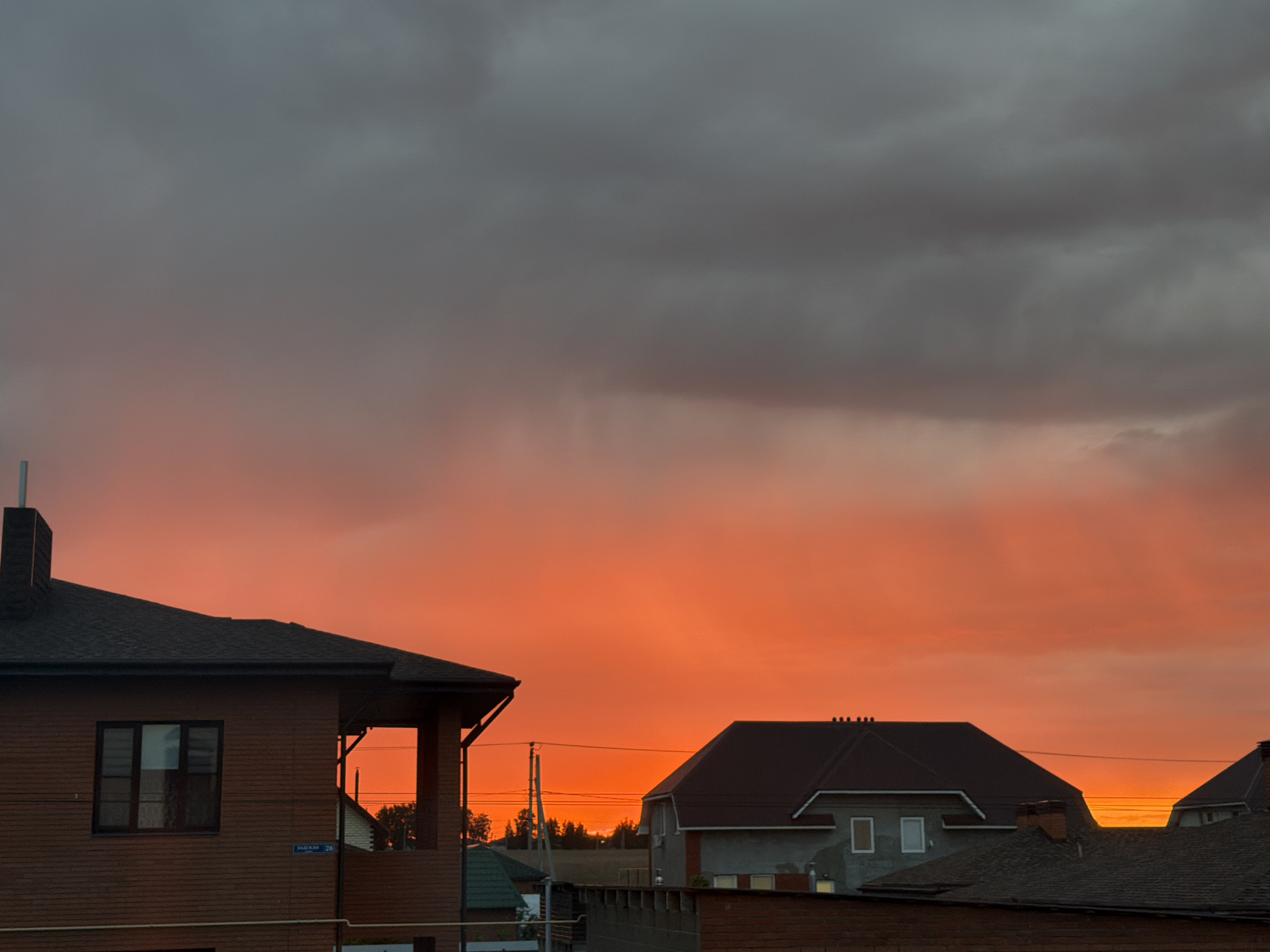 The image depicts a twilight scene in what appears to be a residential neighborhood in Большая Шильна, Russia. Several buildings with dark roofs and brick or stone facades are visible, silhouetted against a vibrant sunset. The sky transitions from a fiery orange near the horizon to dark gray clouds above. Power lines are visible, cutting across the sky in the background. A street sign is attached to one of the buildings on the left. The composition suggests a static view from ground level.
