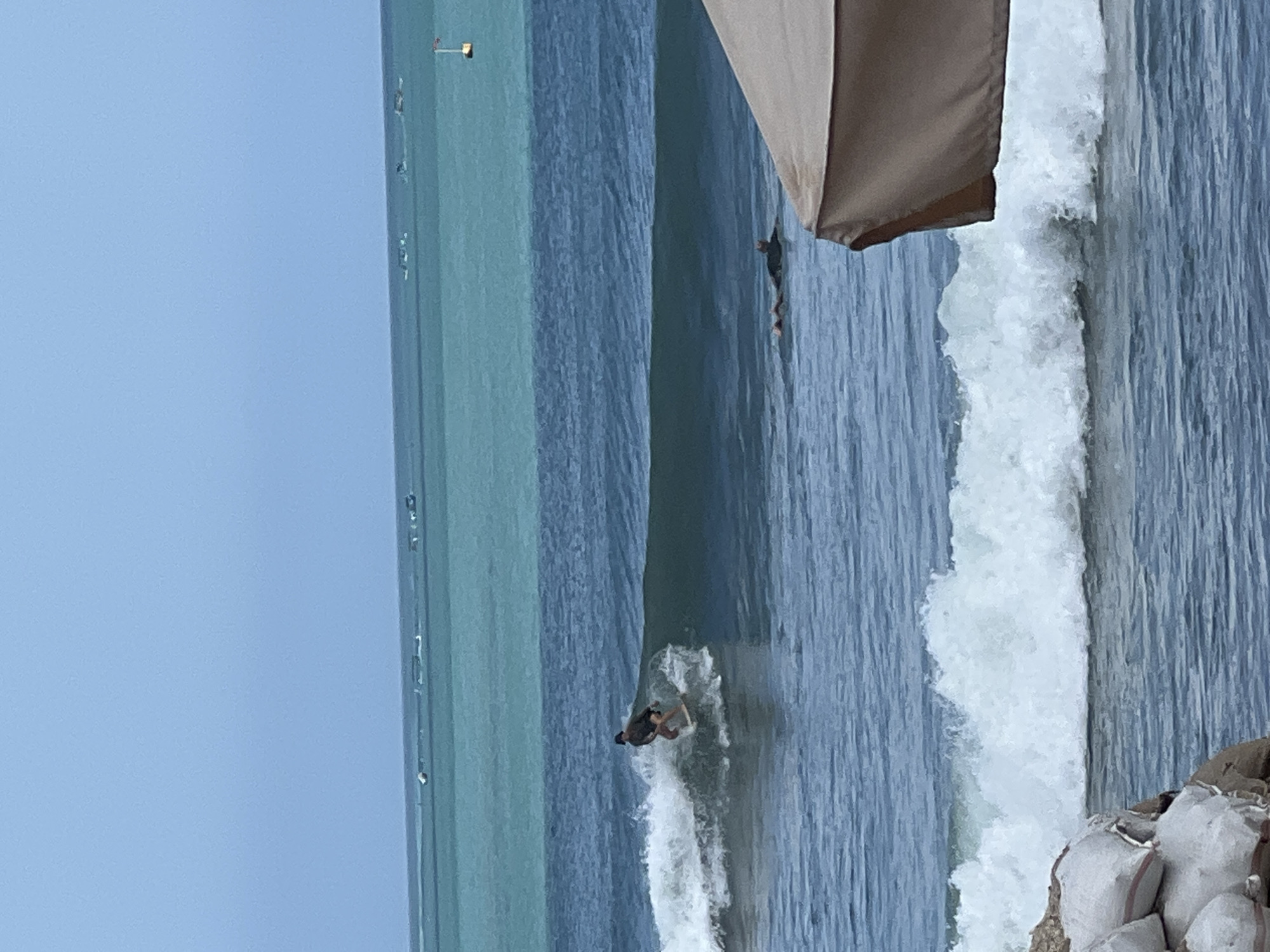 The image captures a segment of the ocean near Kuta, Indonesia, on a cloudy day. The scene is divided into three horizontal bands of water, with the furthest band appearing lighter and the closest bands darker. In the middle band, a surfer is actively riding a wave, leaning to the left on a light-colored surfboard. The surfer is wearing a dark jacket and shorts. Further out in the middle band, another person is lying on their back in the water, facing the sky. The closest water band features a large, foamy wave breaking. In the foreground, the edge of a beige umbrella canopy is visible on the right, and on the left, a pile of sandbags is stacked. In the background, the horizon is dotted with numerous small boats, and a yellow buoy with a red top is visible in the turquoise section of the water.