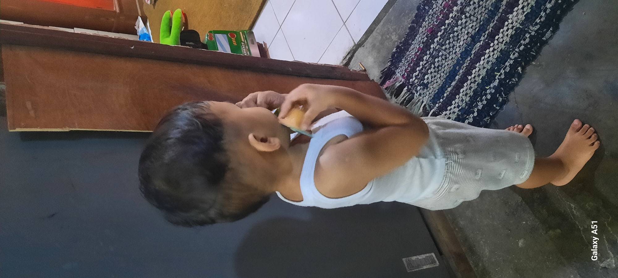 A toddler wearing a white tank top and light-colored shorts is standing on a dark gray floor indoors, near a doorway. The toddler is facing away from the camera, with their right side visible, and is holding a small, light-colored object, possibly food, to their mouth with both hands. A woven rug with dark and light stripes is visible on the floor to the toddler's right. The background includes a dark blue wall to the left and a wooden door frame to the right, which opens to a room with a tiled floor and a green toy. The image is a full-frame shot of the toddler and their immediate surroundings. The location is specified as Jatitengah, Indonesia.