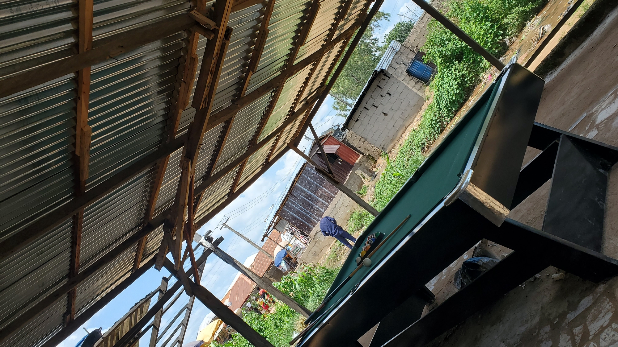 A tilted, full outdoor shot shows a billiards table situated under a corrugated metal roof supported by rough-hewn wooden beams. The pool table, covered in green felt, has balls and cues arranged on it. In the background, a person in a dark blue outfit is seen bending over. Surrounding the pool table and the background are various structures including brick buildings, some with corrugated metal siding, and an assortment of vegetation. A blue barrel is visible near one of the buildings. The scene suggests an informal outdoor entertainment area, potentially in a residential or semi-commercial part of Jalingo, Nigeria. The lighting indicates it is daytime, with the sky visible through gaps in the roofing.