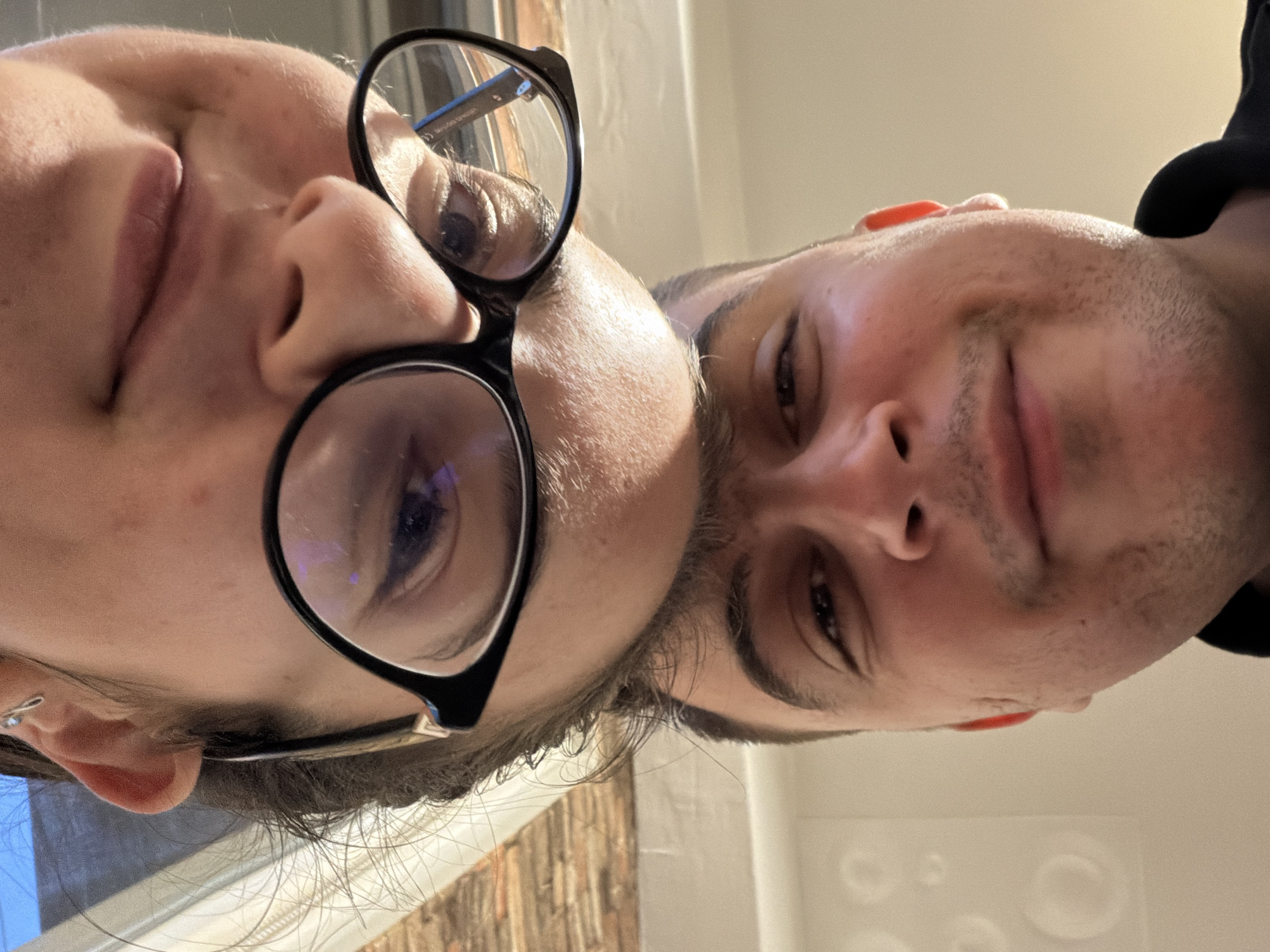 Two individuals pose close together in an indoor setting. The person on the left wears glasses with a thick black frame. The background includes a window and a brick-patterned wall. The location is not discernible as Sofia, Bulgaria.