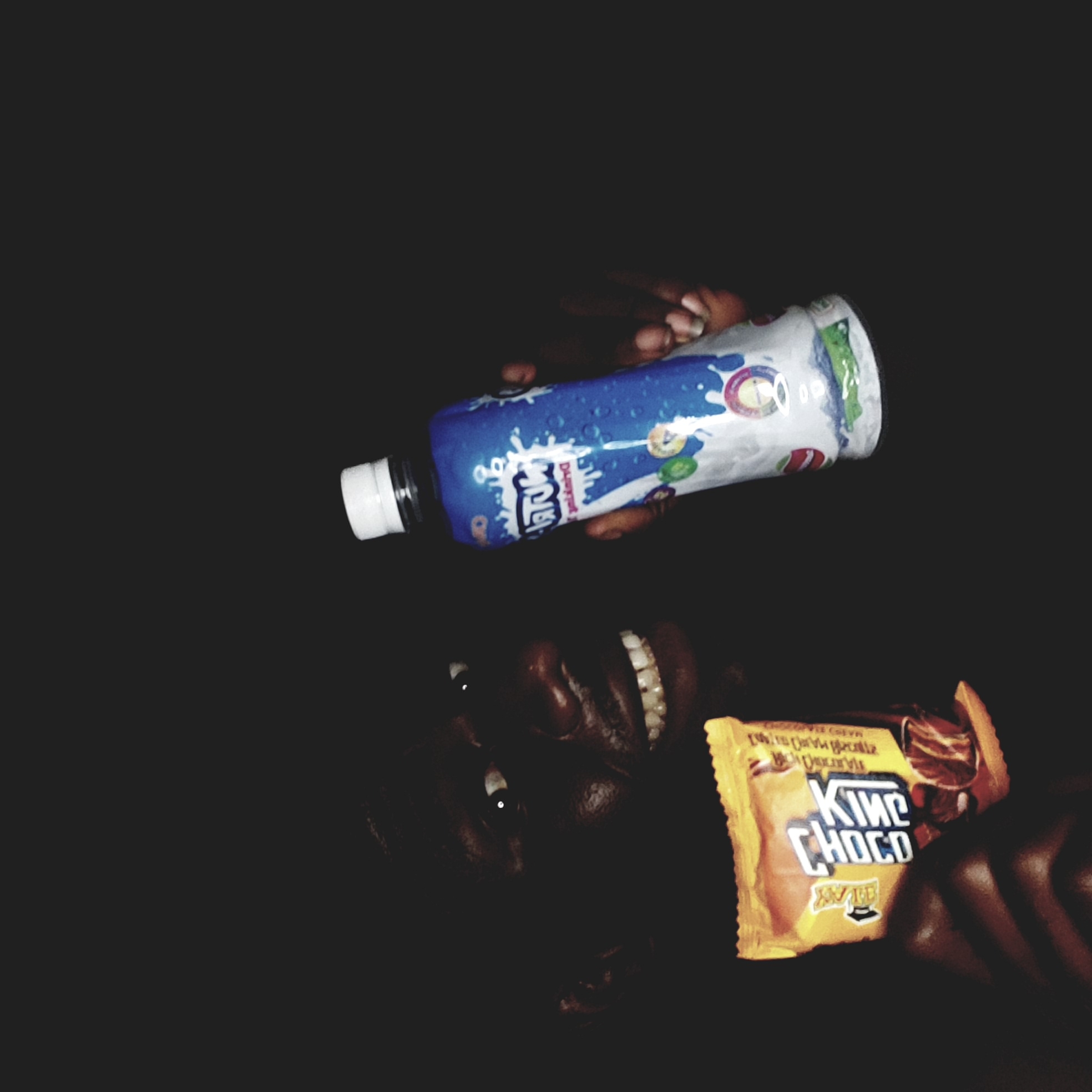A person lies in near darkness, holding a bottle of Yop dairy drink and a King Choco chocolate bar. The exact location within Narayi, Nigeria, cannot be determined from the image.
