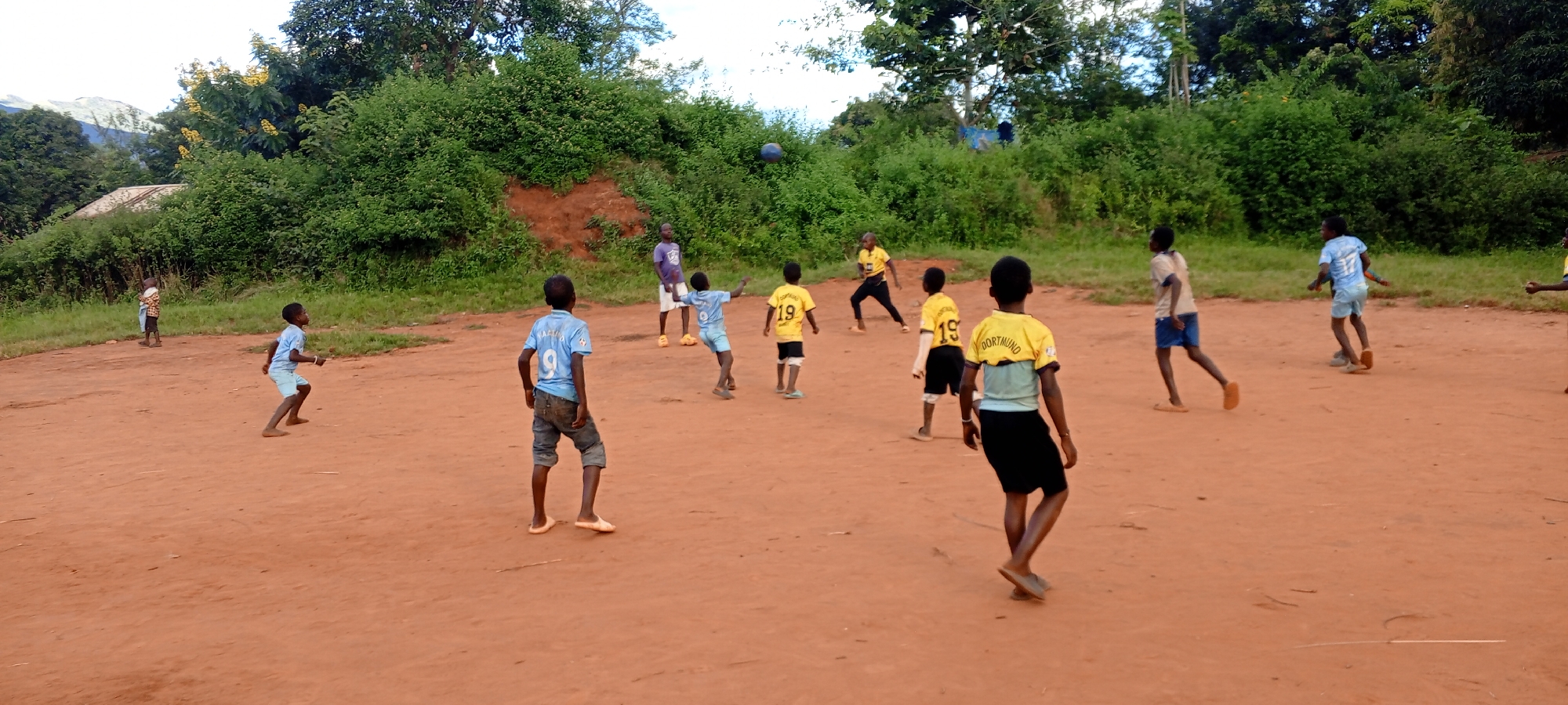A daytime wide shot shows a group of children playing soccer in an open dirt field in Ngalula, Democratic Republic of the Congo. The children, barefoot or wearing simple footwear, are dressed in various sports jerseys and shorts. Several wear yellow jerseys with "DORTMUND 19" on the back, while others are in light blue shirts with numbers. One child is dressed in a purple shirt and white shorts. They are actively engaged in playing, with a dark-colored ball in the air. The field is reddish-brown dirt, with green vegetation and trees in the background. A building is partially visible behind the trees on the left. The sky is overcast.