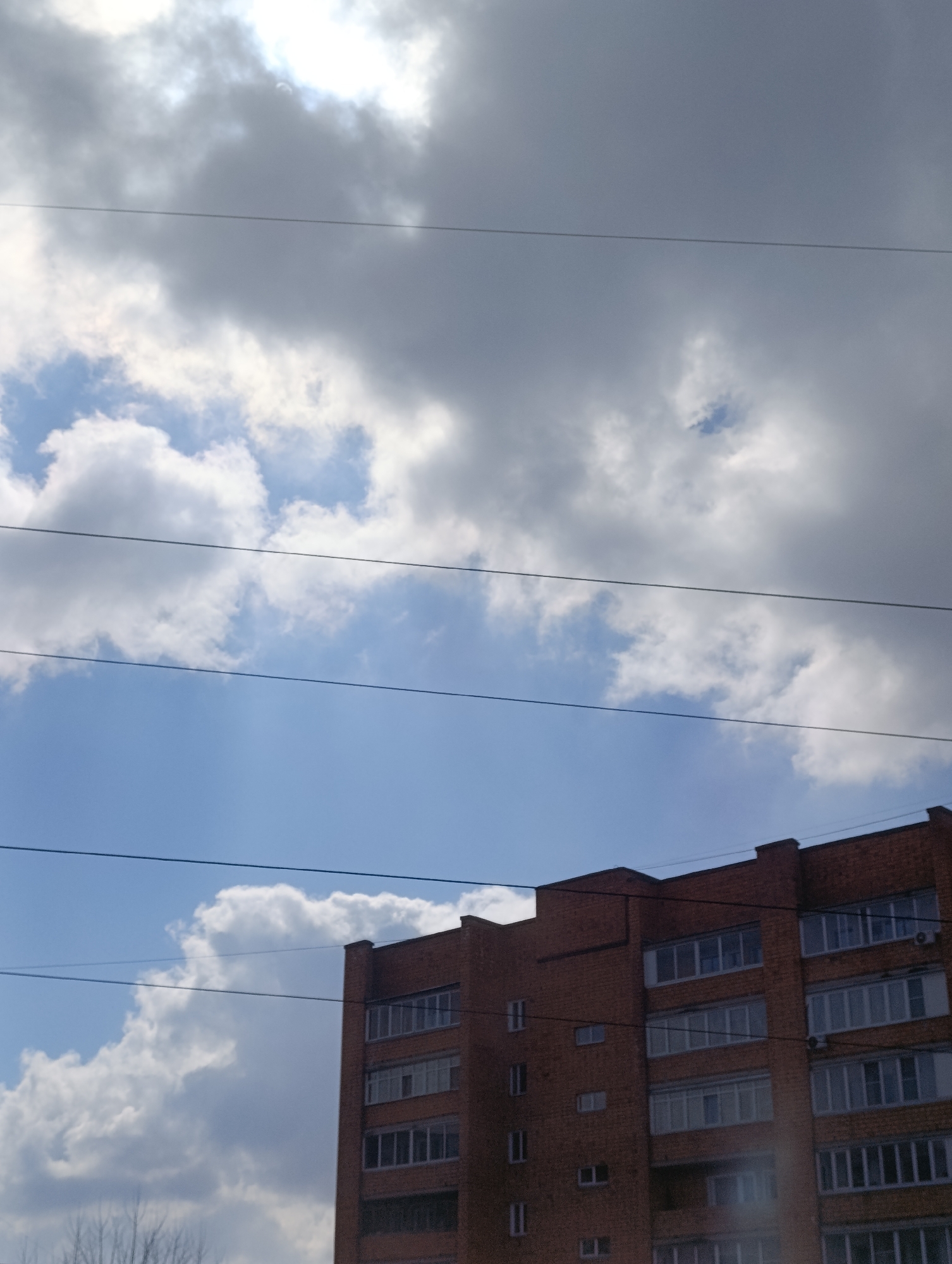 The image shows a brick apartment building under a cloudy sky with some blue patches, potentially located in Krasnoyarsk, Russia. The building appears to be several stories high, with numerous windows. Power lines are visible crisscrossing the sky. The lighting is somewhat diffused due to the cloud cover. There are trees at the base of the building. The sky is primarily light blue and white.