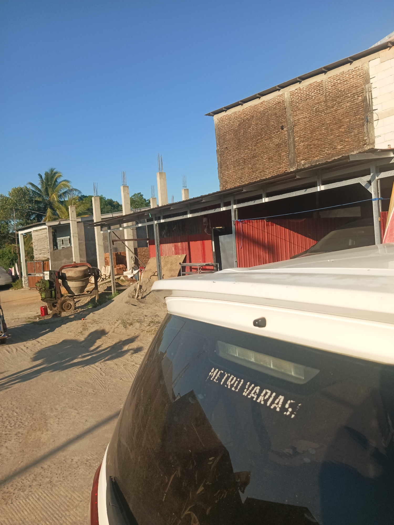 The image depicts a construction site with a building under construction and a vehicle in the foreground. The building has exposed brickwork and concrete pillars, indicating ongoing construction. A cement mixer is visible on the site, along with piles of sand and bricks. The environment appears to be outdoors with a clear blue sky. In the foreground, the rear of a white vehicle is visible, with a logo that appears to read "METRO VARIA S". The presence of construction materials and unfinished structures suggests that this scene is located in a developing area. Based on the provided information, this construction site is located in the city of Pangkajene, Indonesia. The lighting and shadows suggest that the photograph was taken in the late afternoon or early morning.