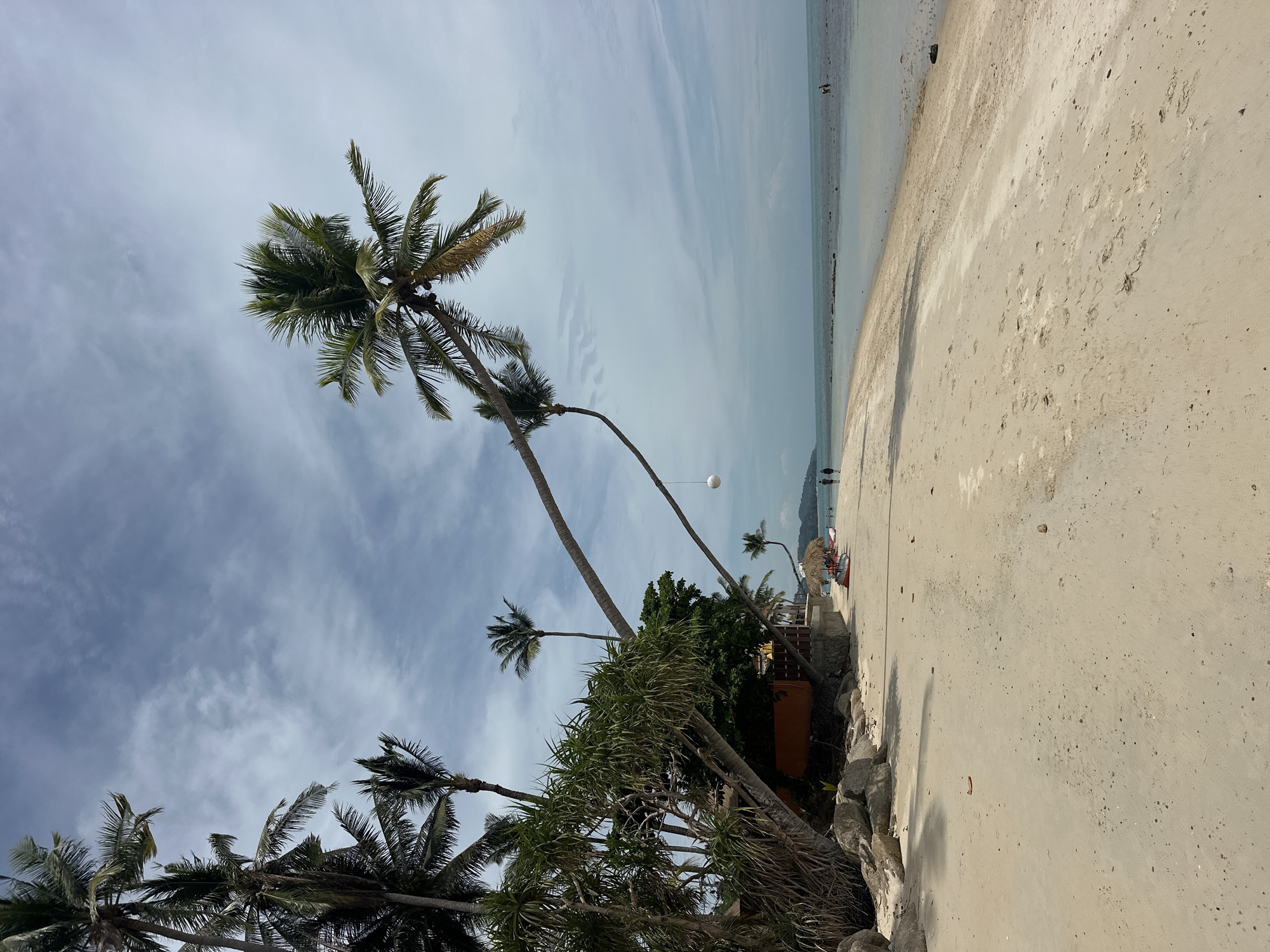 This image depicts a tropical beach scene in the Ko Samui District of Thailand. The foreground is dominated by a wide expanse of light-colored sand, showing subtle textures and scattered small pebbles. To the left, a dense cluster of vegetation, including palm trees with their characteristic fronds, lines the edge of the beach. Some of these palm trees lean dramatically over the sand, with one particularly large palm tree arching across the frame from the left. Beneath the vegetation, a small building with an orange-tiled roof is partially visible, suggesting a resort or beachfront establishment. Further along the beach, a thatched umbrella provides shade over what appear to be two red kayaks.
In the mid-ground, a calm, shallow body of turquoise water meets the shore. The water recedes to reveal a seabed with scattered rocks and sandbanks. A few individuals are visible on the beach and in the water. Two people are walking along the shoreline in the distance, and two others are wading in the shallow water. Another person is visible further out in the water. The sky above is a pale blue, filled with wispy, scattered clouds, indicating a partly cloudy day. The overall atmosphere is serene and idyllic, characteristic of a beach destination.