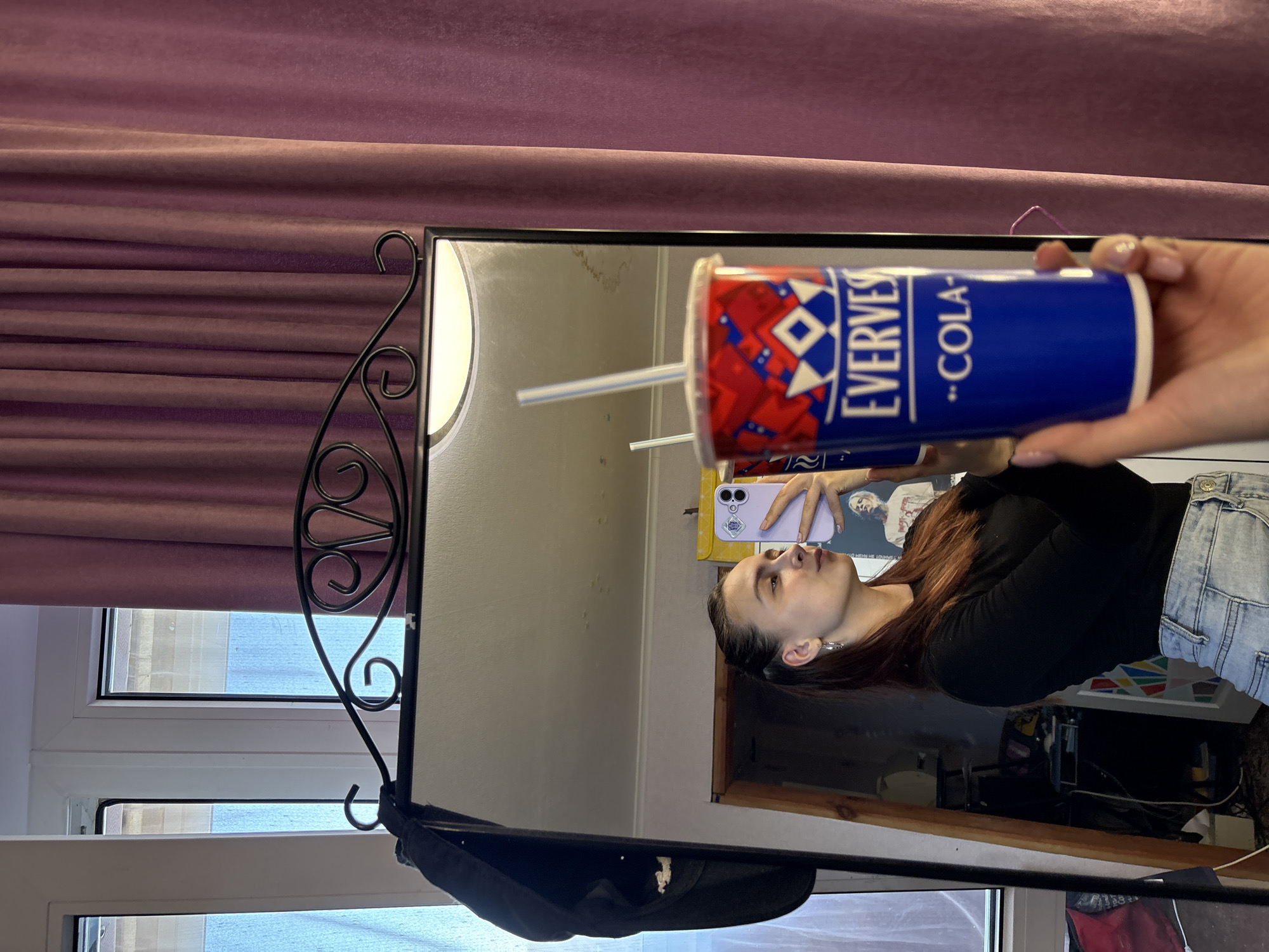 A person holds a "Evervest" Cola cup in front of a mirror. The reflection shows a woman taking a picture with her phone. The window and the reflection of the window suggest the image was taken in a brightly lit area. A black hat hangs on the mirror stand. The location in Прикамский, Russia is not deducible from the provided image.