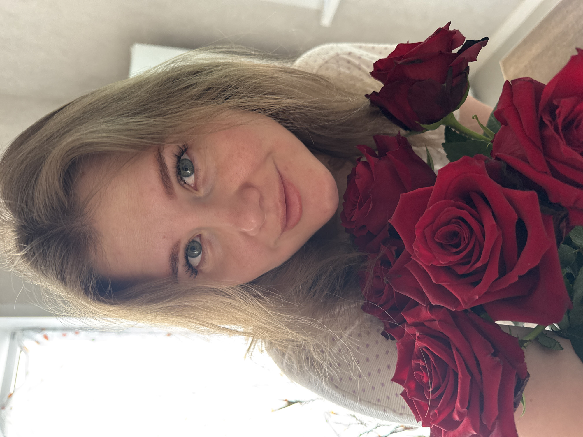 A woman with light blond hair is smiling and holding a bouquet of red roses. The photo appears to be taken indoors near a window with light coming through. The background is blurred, but it looks like a wall with a window and some furnishings. The location is near Gornyi (Dvurechenskoe S/P), Russia, though this cannot be definitively determined from the image alone.
