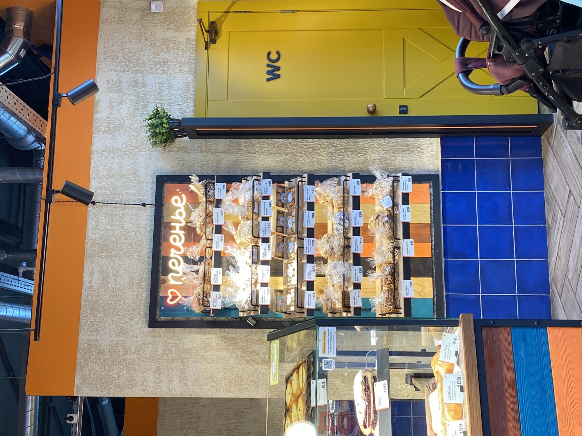 This image depicts a bakery or confectionery shop in Murino, Russia. The shop features a display of various types of cookies and pastries, each labeled with its price in Russian currency. The cookies are packaged in clear plastic and arranged on a metal rack behind a glass display case. Above the display, there is a sign with the word "печенье" (cookies) written in neon letters. The background includes a bright yellow door with the letters "WC," indicating a restroom. The scene is well-lit, with artificial lighting highlighting the displayed goods. The style of the shop suggests a modern design, with a mix of rustic and industrial elements. A baby stroller is partially visible in the background, suggesting a possible location near a family-friendly area.