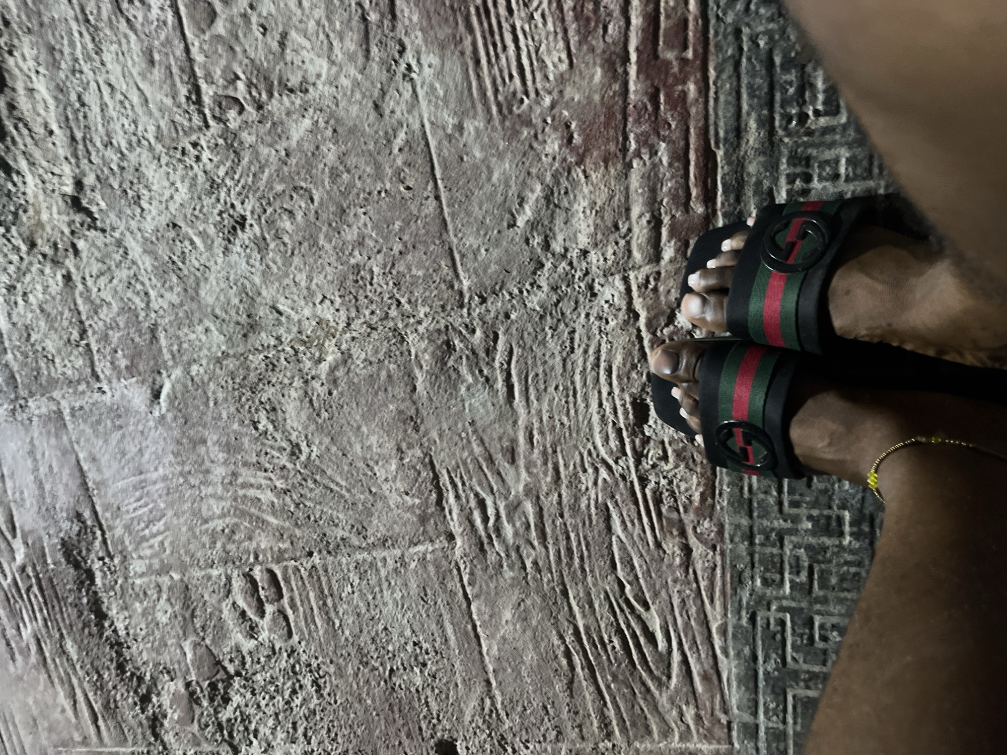 The image shows a close-up view of a person's feet wearing Gucci slides on a textured, patterned surface that resembles a street or pathway. The surface has a weathered, stone-like appearance with visible lines and indentations, possibly suggesting a cobblestone or carved concrete design. The lighting appears to be low, creating shadows that highlight the texture of the ground. One foot is adorned with a delicate gold bracelet with small yellow beads. The overall impression is a candid shot of someone's feet in an outdoor urban setting. Without further information or context, pinpointing the exact location within Abuja, Nigeria, is not possible from the visual elements alone.