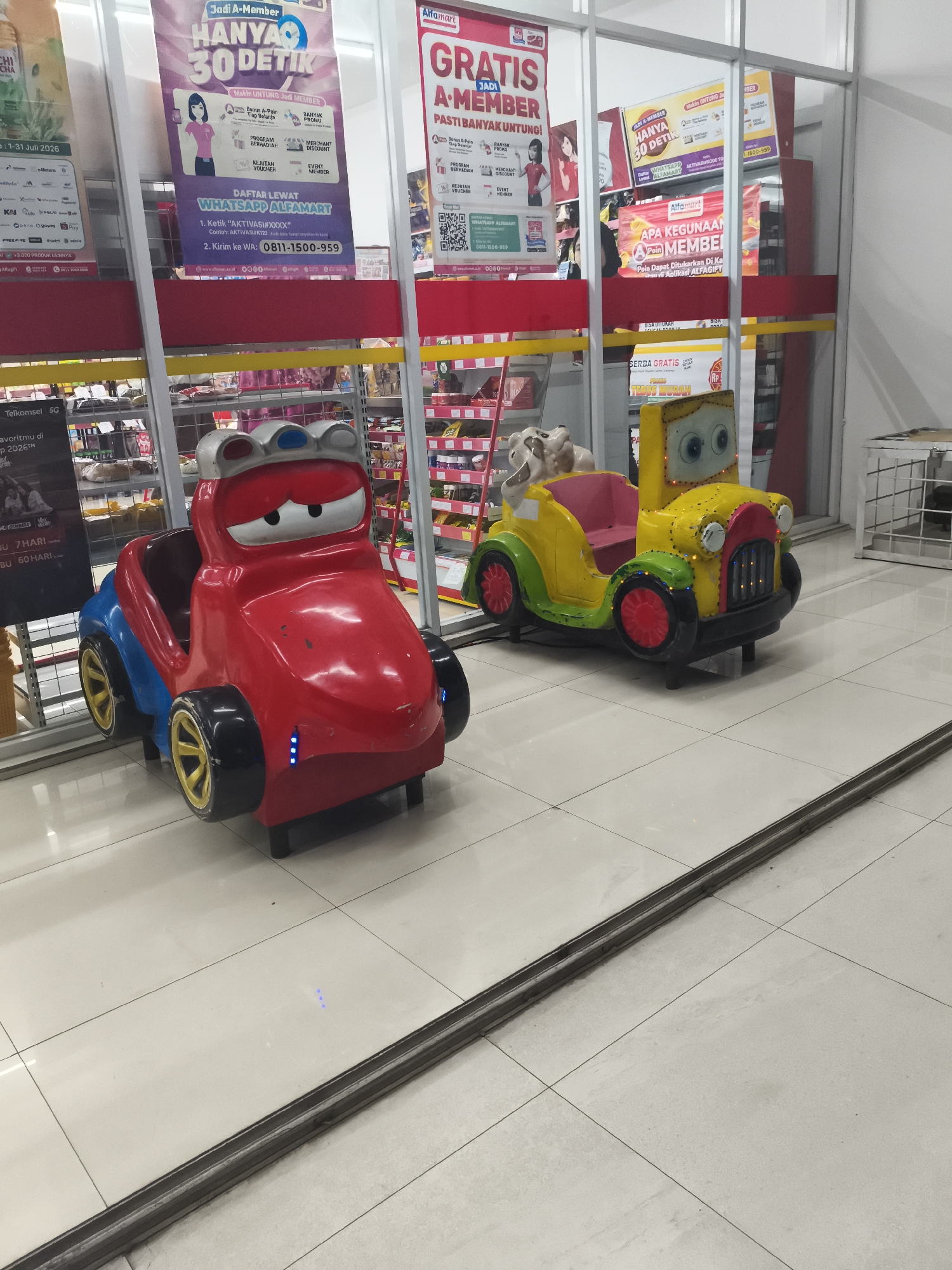 In Depok, Indonesia, two brightly colored children's ride-on toys are positioned on a tiled floor inside a commercial establishment, likely a convenience store, given the signage visible in the background. To the left is a red and blue toy car with a cartoonish face, featuring large white eyes with blue pupils, a metallic silver and blue crown-like accessory on its head, and yellow rims on its wheels. To the right is a yellow toy vehicle with green accents and red wheels, resembling a vintage car with a smiling face depicted by large blue eyes and a black grille. The background reveals shelves stocked with merchandise, and large promotional banners are visible, including one for "Alfamart" promoting membership benefits and another for "Jadi A-Member Hanya 30 Detik". The floor is a light-colored tile with dark grout lines, and a metallic strip runs horizontally across the foreground. The overall lighting suggests an indoor environment with artificial illumination.
