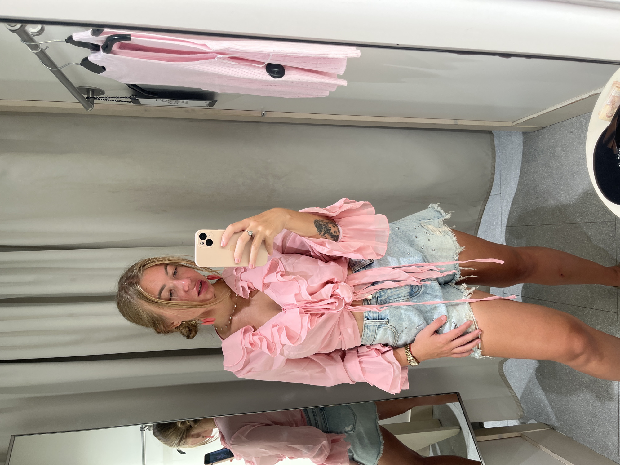 A young adult female is standing inside a retail changing room, taking a selfie with a light-colored smartphone held in her right hand. She has blonde hair styled in a bun with strands loose around her face. Her expression shows a slight pout as she looks towards the phone. She is wearing a sheer, light pink crop top with a deep V-neck, featuring multiple ruffles along the neckline and cuffs, and tied at the front waist. Her lower garment consists of light blue, distressed denim shorts with frayed hems and small, scattered embellishments. She wears reddish-pink dangling earrings, a delicate beaded necklace, a gold-toned watch on her left wrist, and a ring with a clear stone on her right ring finger. A tattoo depicting stylized faces is visible on her right forearm.
To her left, a large rectangular mirror reflects her left side, showing her profile and the back of her shorts appearing as a denim mini-skirt due to the angle. Behind the reflection, a light-colored curtain is partially visible. The changing room walls are light neutral in color, possibly fabric-lined, and the floor is a light grey mottled tile. On the right wall, a silver-toned metal clothing rack holds several garments on hangers, including a pink ribbed top with a black security tag attached. In the bottom right foreground, a low, round white table or stool is partially visible, topped with a black object (possibly a bag or hat) and a folded item. The scene is an unspecified commercial establishment located in the city of บ้านบางรักษ์, Thailand.