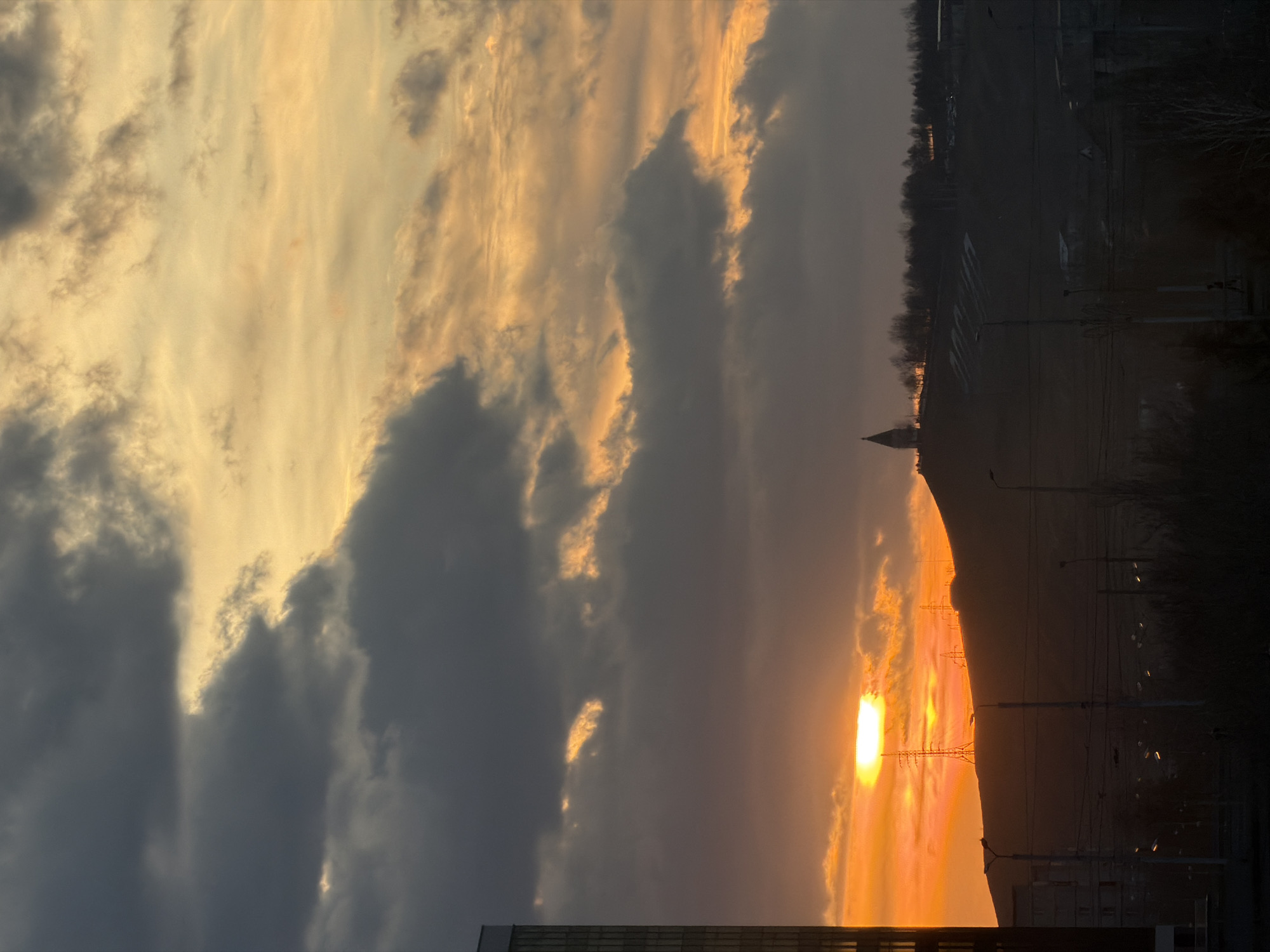 The image captures a sunset in Krasnoyarsk, Russia. The sun is low on the horizon, emitting a bright orange glow. Overlapping clouds are illuminated by the sunlight, creating a dramatic effect. In the foreground, there are buildings with rectangular shapes and structures silhouetted against the sun. There is a hill on the right side of the image and electrical infrastructure and streetlights. The upper part of the image shows a dark skyline with some trees.