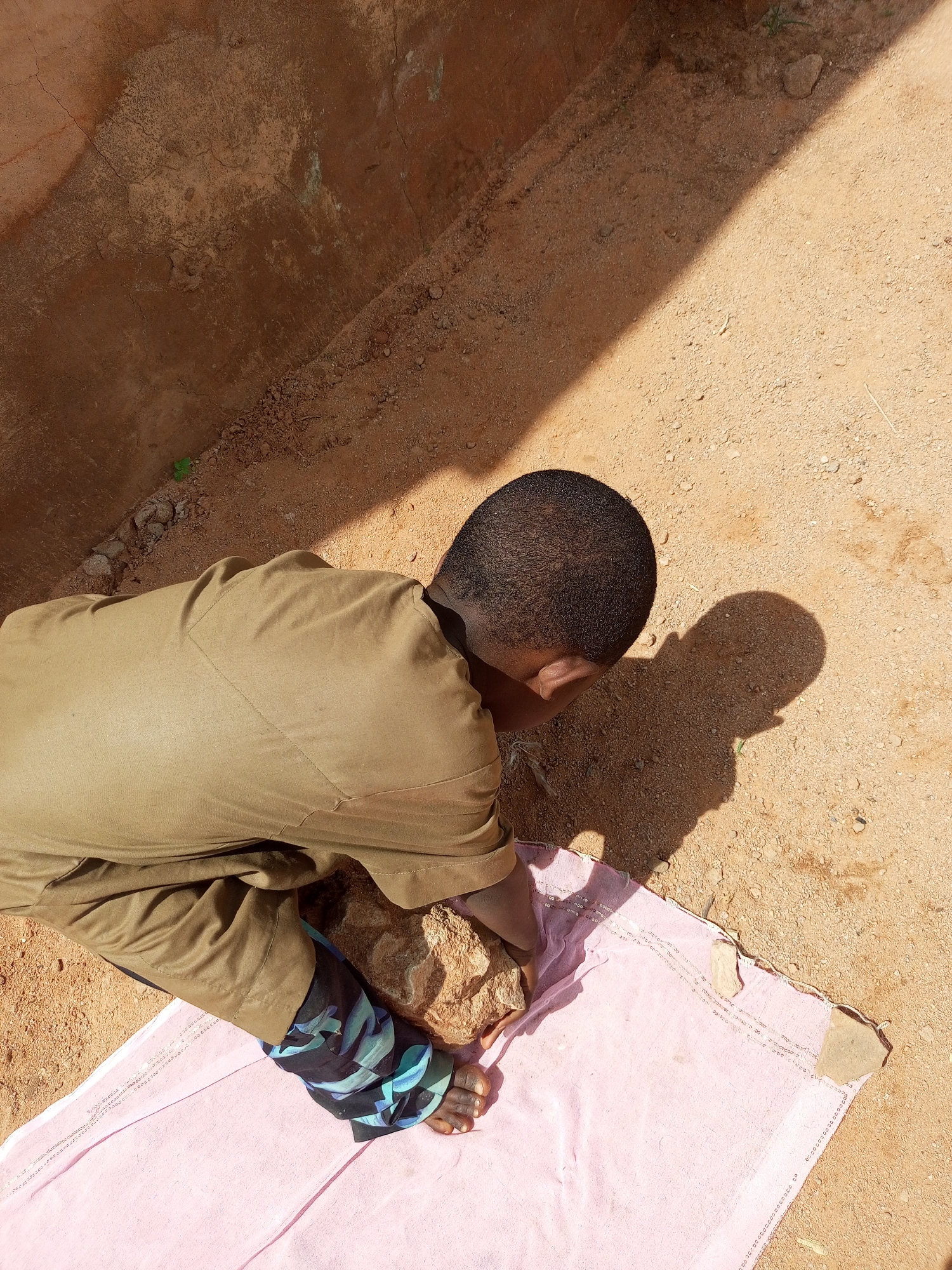 A person wearing a brown tunic and patterned leggings is crouched on a pink cloth on sandy ground. The person is holding a rock in their hands and appears to be examining it. A shadow of the person is cast on the ground beside them. The background consists of a rough, reddish-brown wall and more sandy ground. The scene is lit by sunlight, creating strong shadows. This appears to be an outdoor scene in Gombe, Nigeria.
