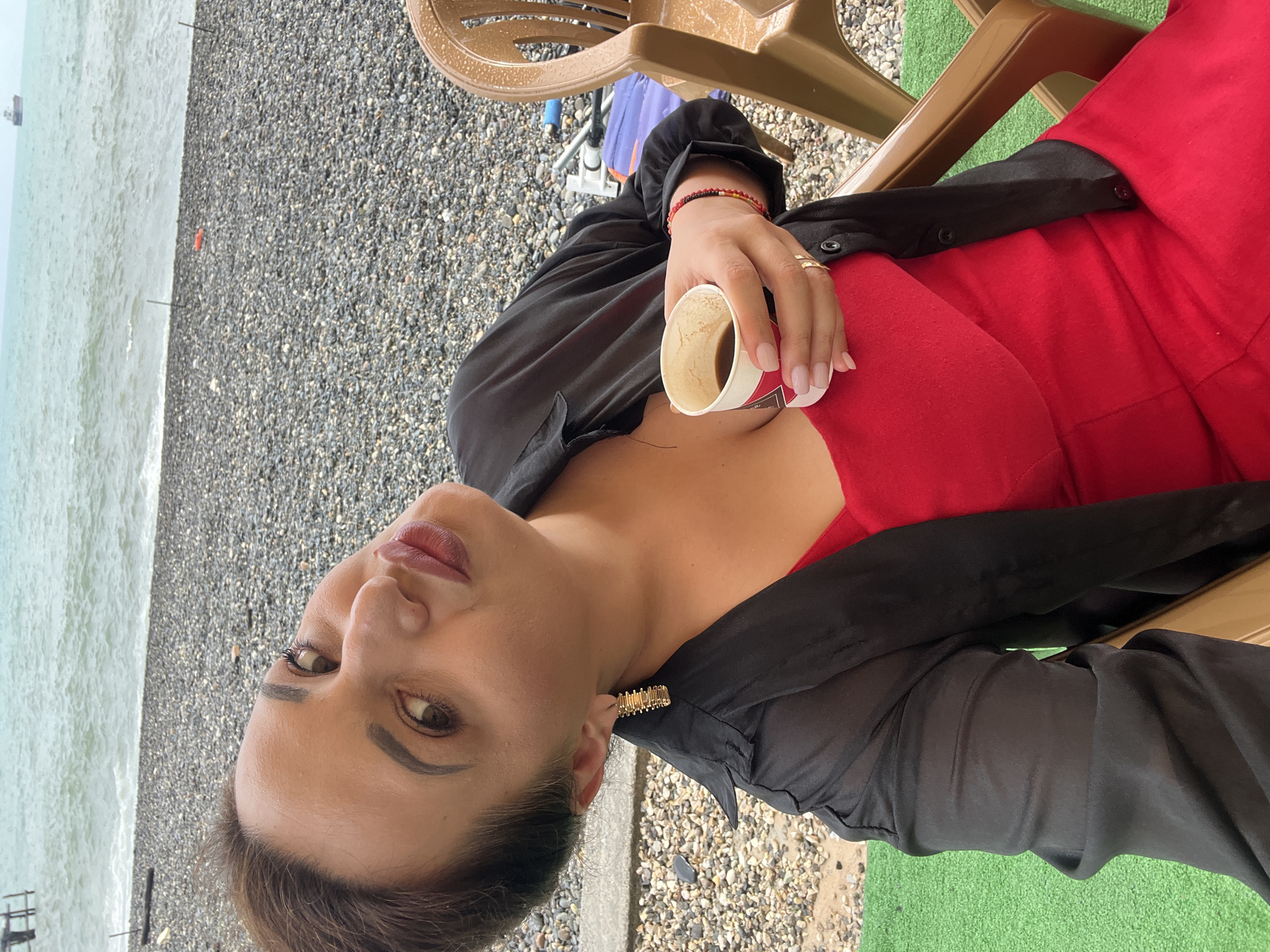 A woman with dark hair pulled back sits on a chair on a pebbled beach with the sea in the background. She is wearing a red dress and a sheer black open-front shirt. In her right hand, she holds a disposable coffee cup filled with coffee. Her left arm rests on her lap, and she is looking towards the camera with a slightly tilted head, her lips pursed. She is wearing a gold earring in her left ear and a red and yellow beaded bracelet on her right wrist. The environment suggests a casual outdoor setting, possibly a cafe or seating area on the beach. The scene is set in the city of Sokhumi, Abkhazia.