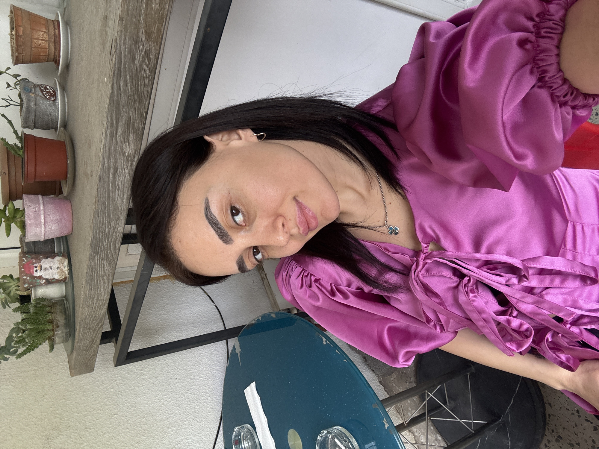 A woman is facing the camera, wearing a purple satin dress. She has dark hair, fair skin, and prominent eyebrows. She is wearing a silver necklace and small hoop earrings.
Behind her, there is a shelf with potted plants and a table with a blue surface. The background includes a white wall and a window.
The exact location is not discernible from the image, but given the prompt, it is assumed to be in Tbilisi, Georgia.